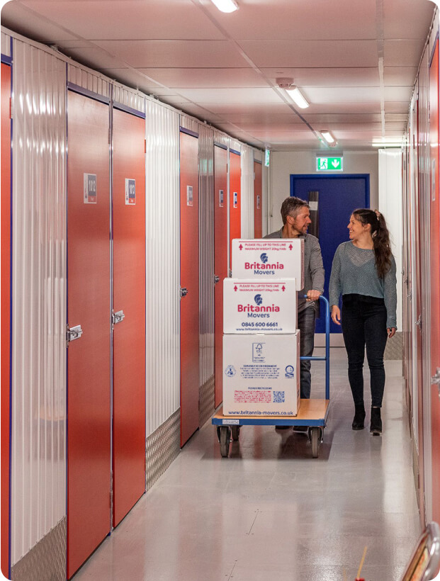 A man and a woman are pushing a dolly with three stacked moving boxes through a hallway of a storage facility. The hallway has red doors on both sides, and a green exit sign is visible at the end. The moving boxes are labeled "Britannia Movers.