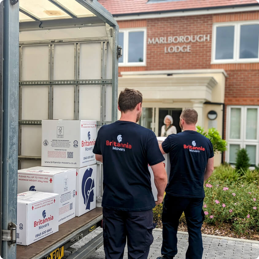 Two movers from Britannia Movers are unloading cardboard boxes from a truck parked outside a building labeled Marlborough Lodge. Both movers are wearing black shirts with the company's logo on the back. The scene is set in a residential area with shrubbery around.