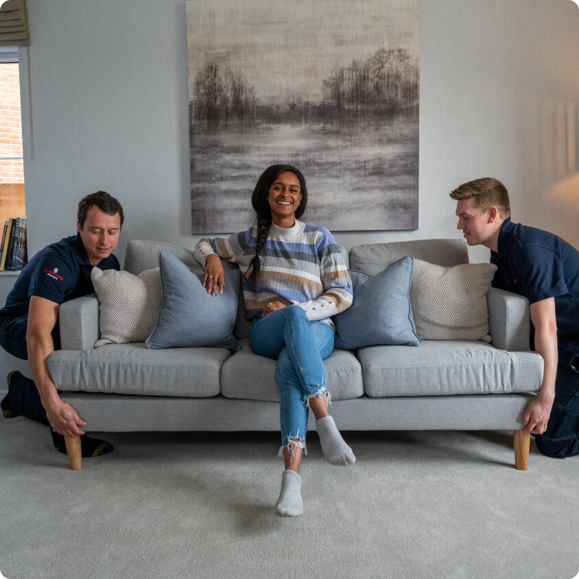 Two movers in uniforms are lifting a gray couch with a smiling woman sitting in the middle of it. The room has light-colored walls and carpet, and a large, framed painting of a nature scene hangs on the wall behind them.