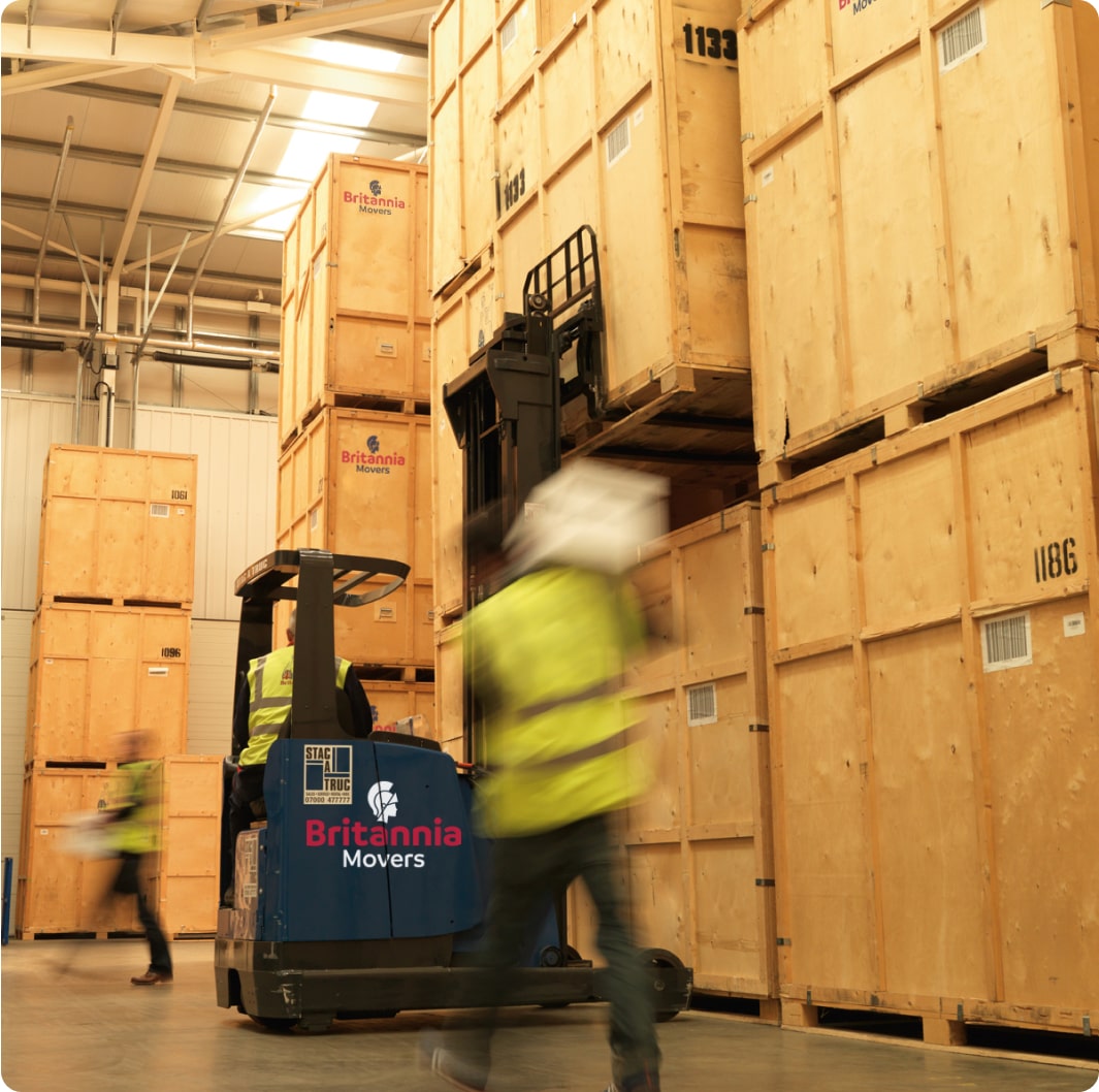 Two workers in yellow safety vests manage large wooden crates in a warehouse with a "Britannia Movers" logo. One operates a forklift, while the other attends to inventory. The scene emphasizes logistics, storage, and careful handling of goods.