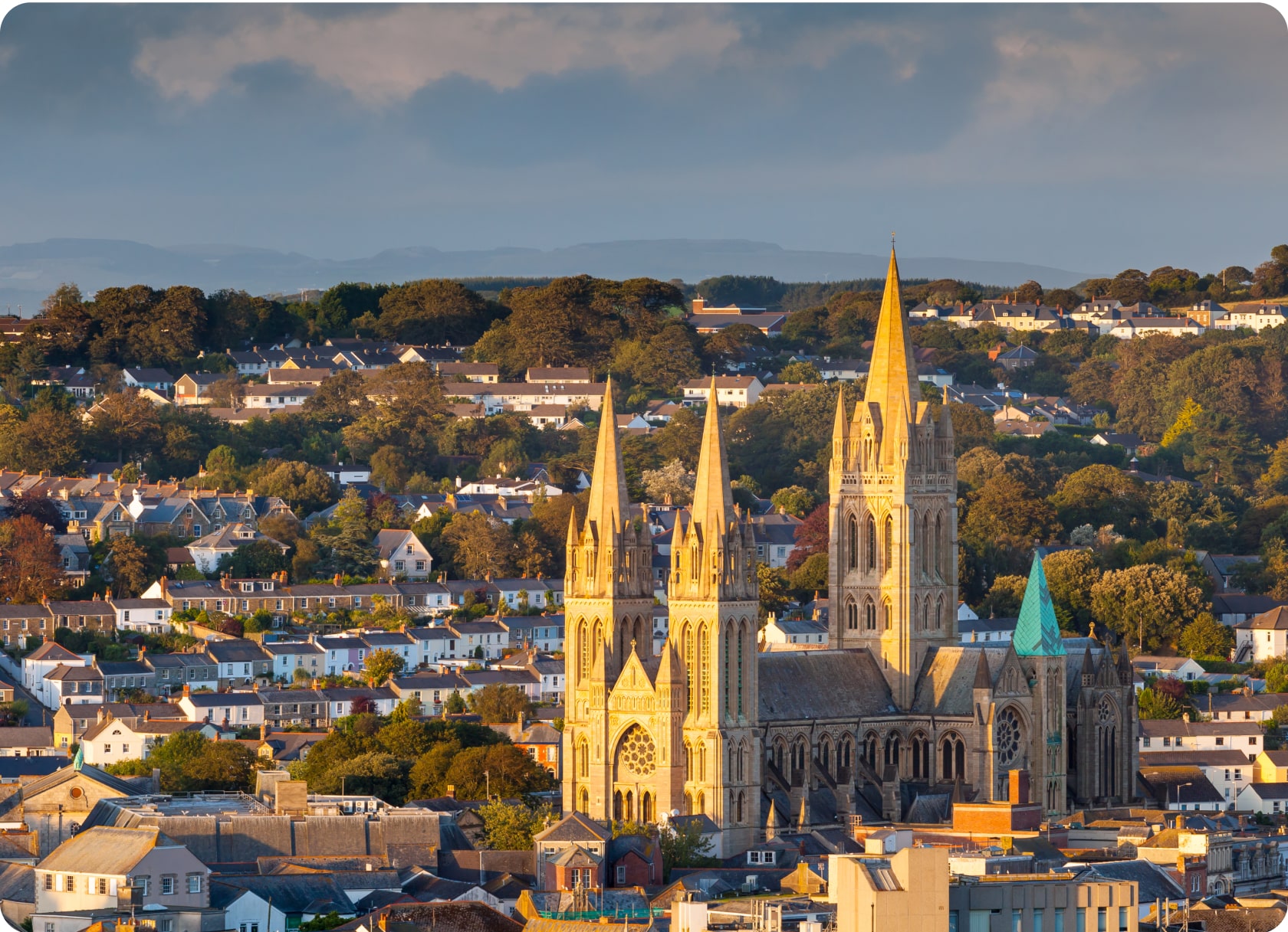 An aerial view of a historic town with a prominent cathedral featuring multiple spires in the foreground. The town is surrounded by lush green hills and a number of houses and trees can be seen throughout the area. The sky is partly cloudy.