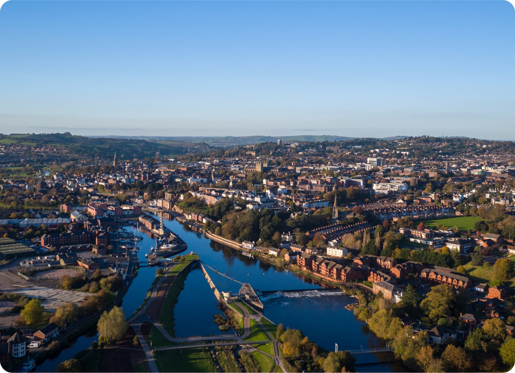 Aerial view of a city with a river running through it, surrounded by green fields and residential areas. The sky is clear and blue, and various buildings and structures are visible along the riverbanks. The landscape extends into the distance with rolling hills.