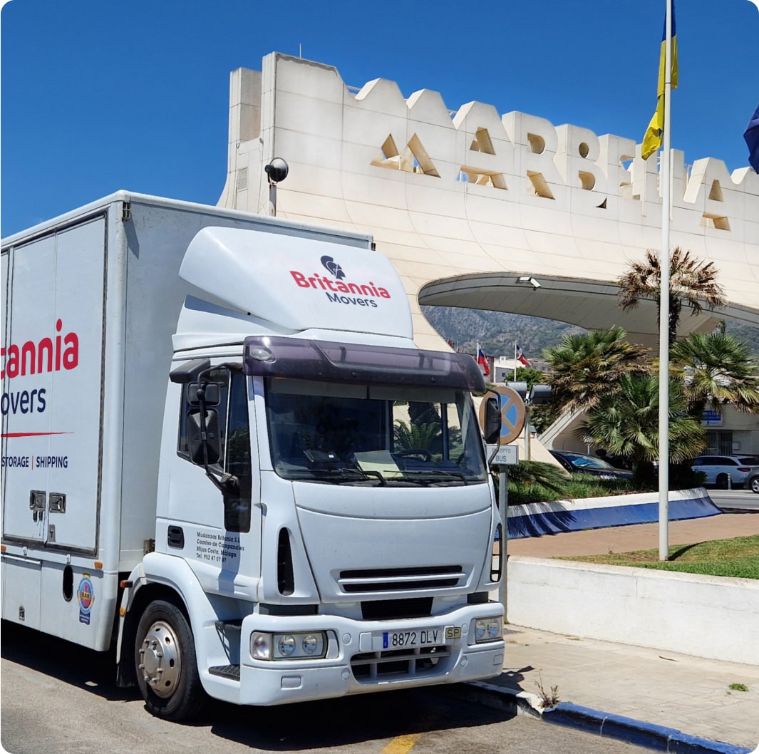 A white Britannia Movers truck is parked in front of a large, modern building with the sign "Marbella" on it. The sky is clear and blue, and several flags are flying next to the building. The scene suggests a sunny day in Marbella.