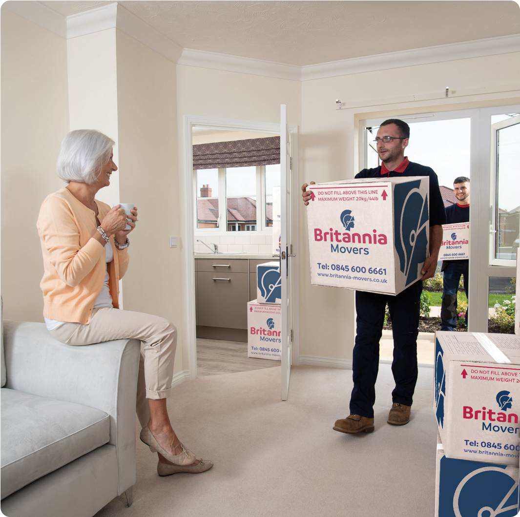 An elderly woman holding a cup sits on a sofa in a living room. Two movers, each carrying a box with the label "Britannia Movers," are in the process of moving items into the room. Another mover stands outside, ready to bring in more boxes.