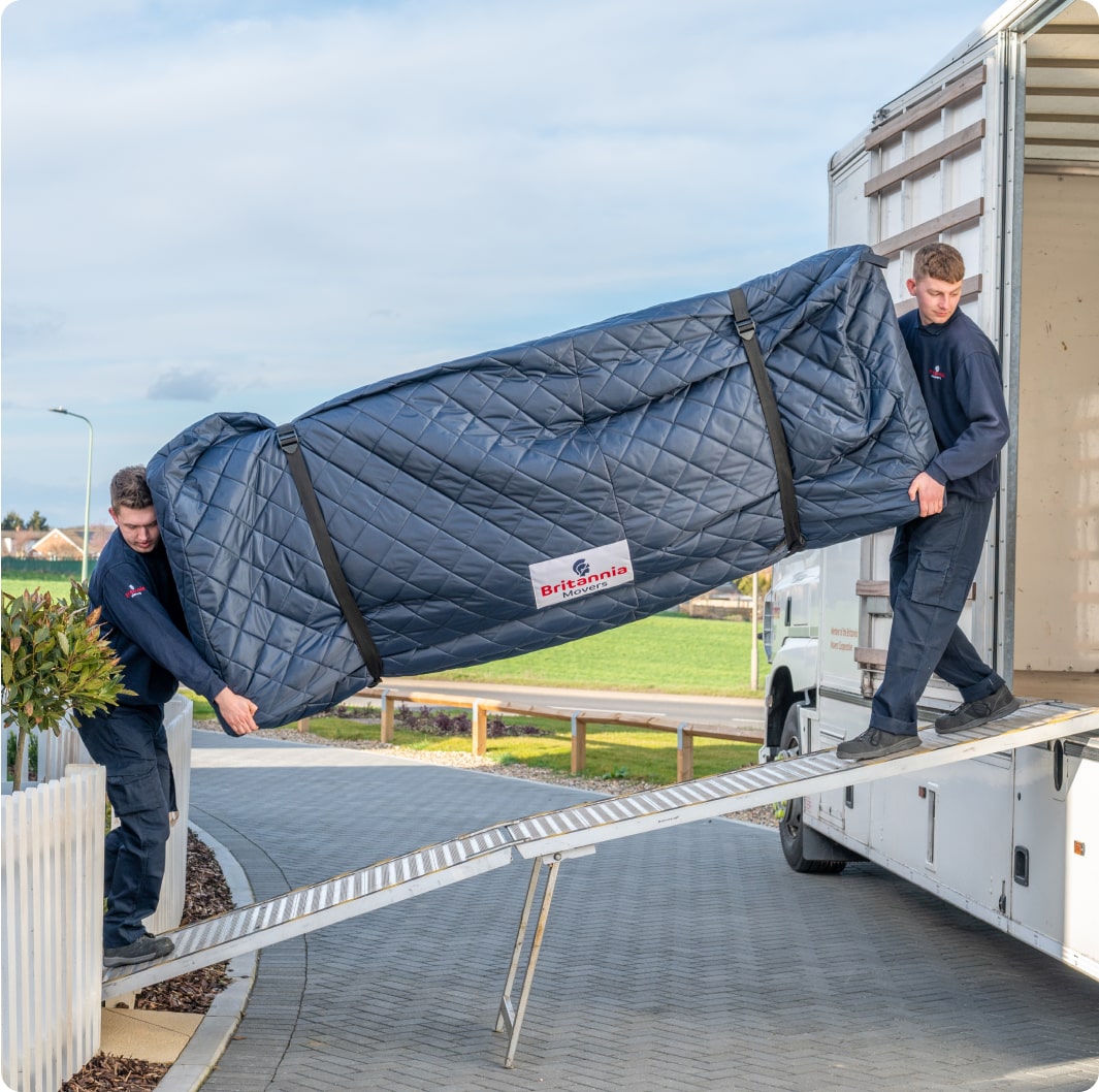 Two movers in blue uniforms carefully carry a large, quilted, navy blue furniture piece wrapped in protective padding up a ramp into a moving truck. A patch on the padding reads "Britannia." A suburban neighborhood is in the background.