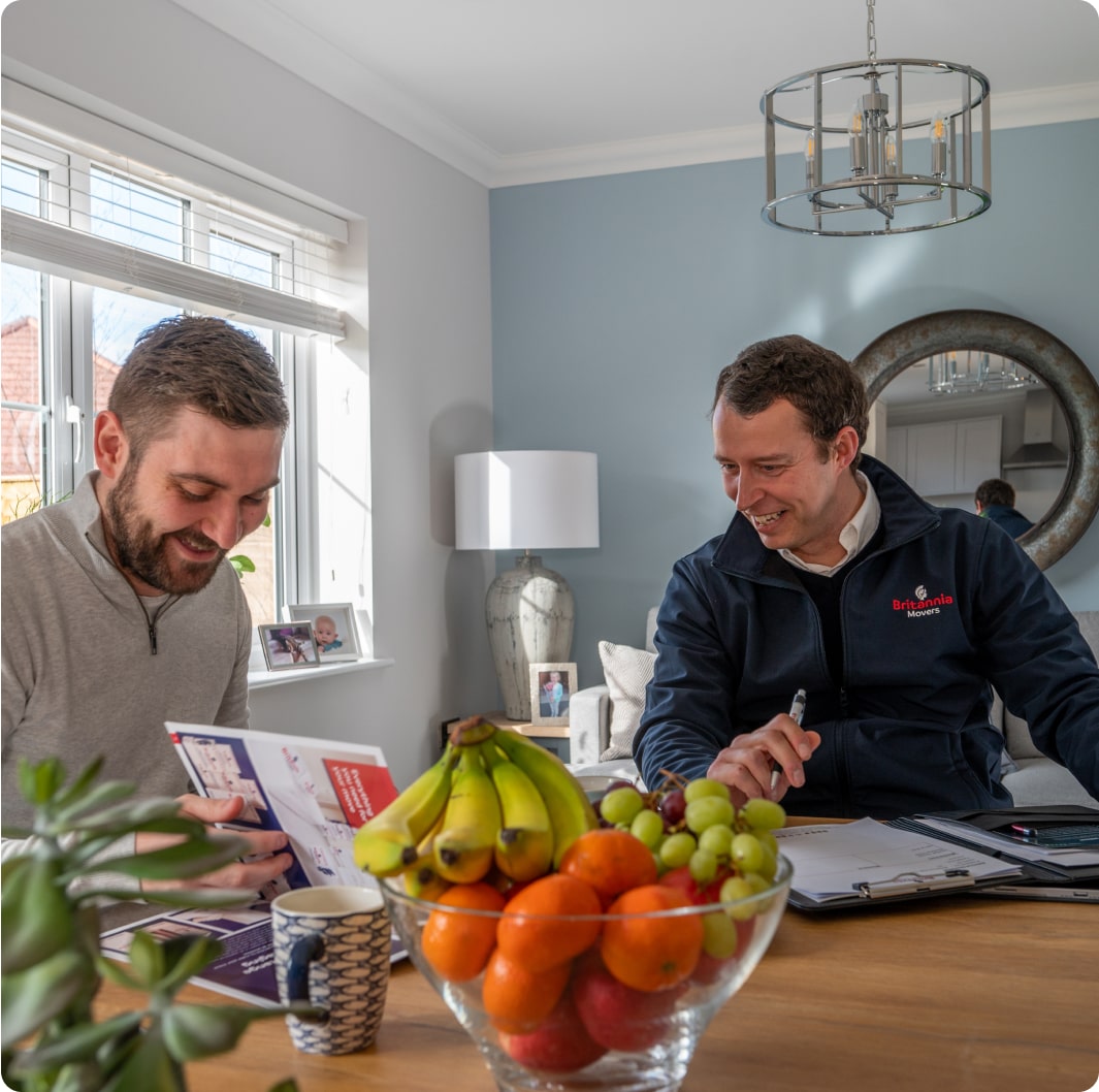 Two men sitting at a wooden table in a modern, well-lit room. They are engaged in discussion over documents. A bowl of colorful fruit is in the foreground. The man on the left wears a light grey shirt, while the man on the right sports a dark jacket.