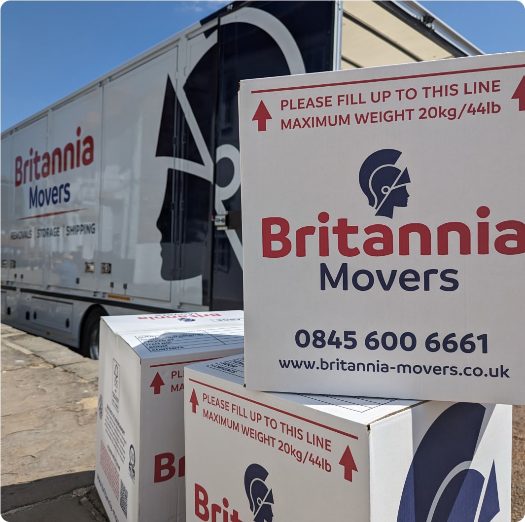 Several white moving boxes with red text and blue logos from "Britannia Movers" are stacked in front of a partially visible moving truck bearing the same branding. The text on the boxes indicates a maximum weight limit and contact information including a phone number and website.