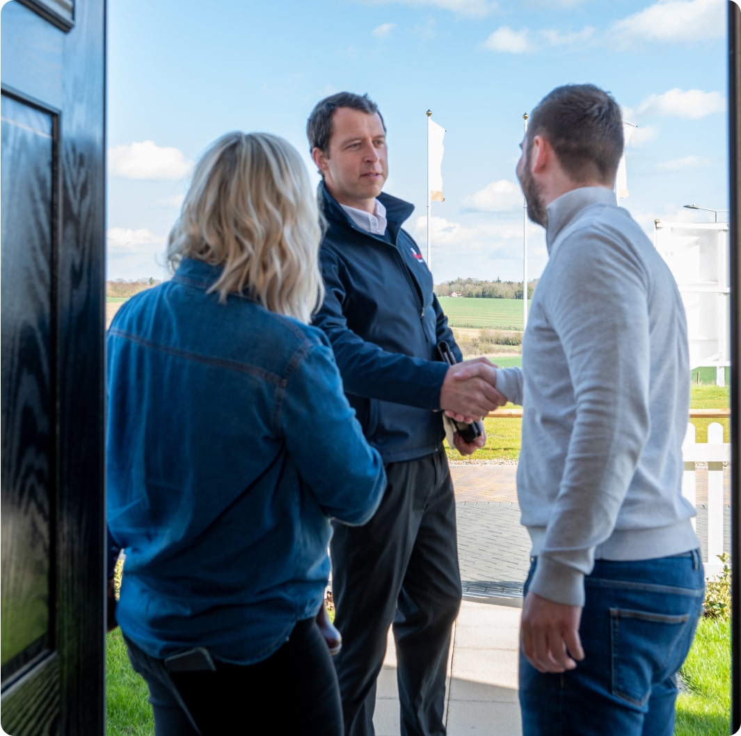 Three individuals are conversing outside: a man and woman stand facing another man. The man and woman with their backs to the camera stand near an open door while the man facing the camera extends his hand for a handshake with the other man. Countryside view in the background.