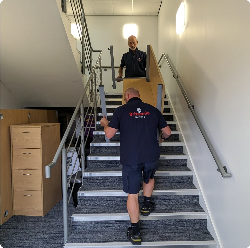 Two movers are carrying a large piece of wooden furniture up a carpeted staircase in an office building. The mover at the bottom is wearing a "Britannia Movers" shirt. They are halfway up the stairs, with one mover on each end of the furniture.