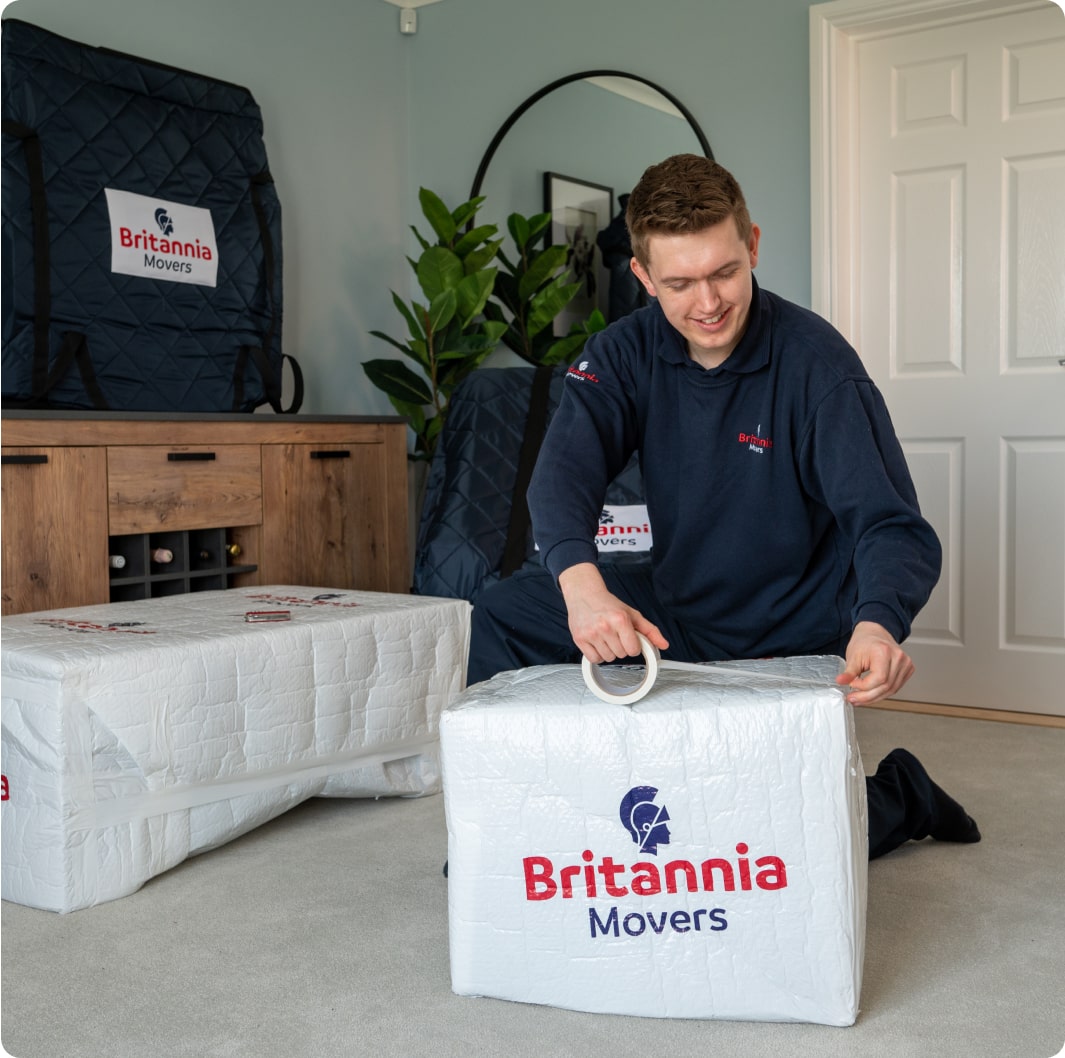 packing-services-img1-min A person wearing a blue uniform is taping a box labeled "Britannia Movers" in a room with a wooden sideboard and a large potted plant. Another box and padded moving covers are visible in the background.