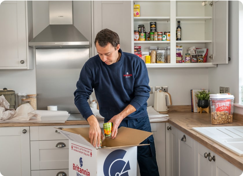 A person is packing canned food into a cardboard box labeled "Britannia" in a modern kitchen. The kitchen cabinets are open, revealing various pantry items. A container of strawberry crisps is on the counter beside the box.