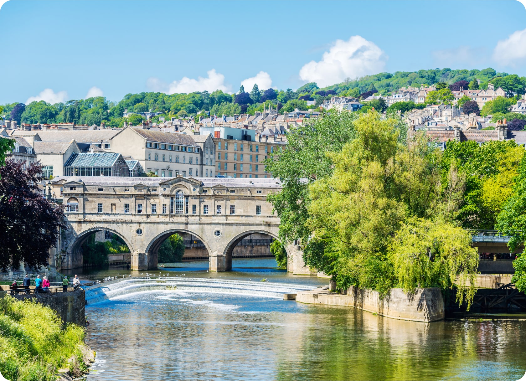 Scenic view of Pulteney Bridge in Bath, England, with its arches spanning the River Avon. Lush trees and vibrant greenery surround the river, while historic buildings are visible in the background. Clear blue sky with scattered clouds overhead.