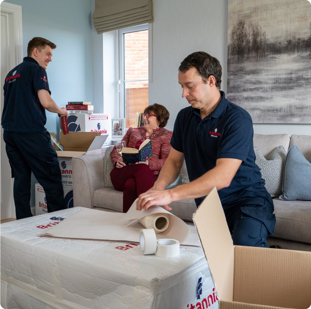 Two movers in blue uniforms are packing boxes in a living room. One is wrapping an item on a table, while the other is carrying a box. An elderly woman with glasses and a red sweater is sitting on the couch, smiling and holding a book, watching them work.