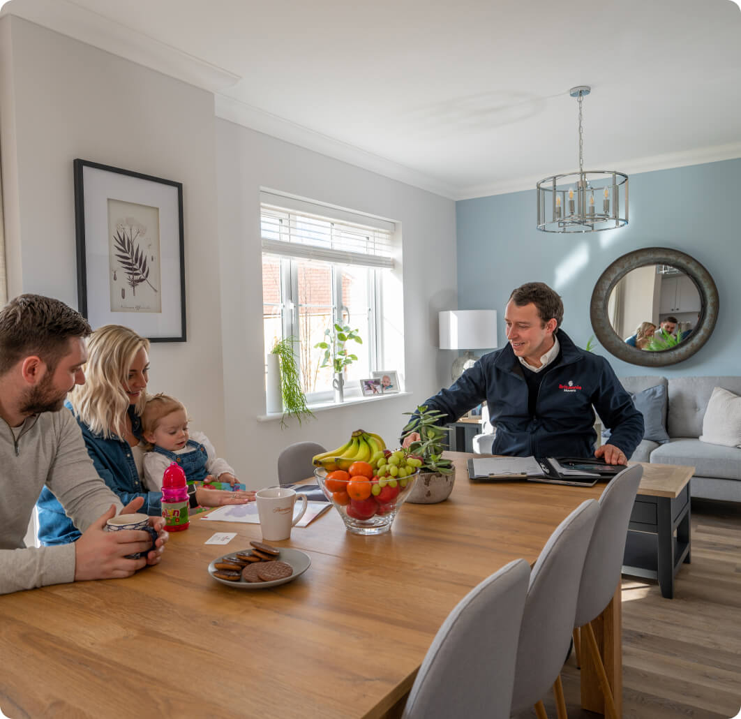 A family sits at a wooden dining table in a bright, modern living room with light blue walls. A man in a jacket, sitting across from them, is showing them paperwork. Fresh fruits, cookies, and a drink are on the table. A couch and a mirror are in the background.