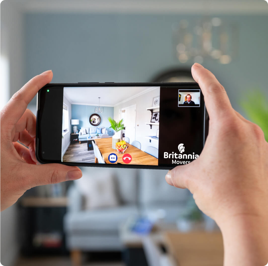 A person holds a smartphone horizontally, conducting a video call via an app with the branding "Britannia Movers" visible. The smartphone screen shows a clear image of a dining room with light blue walls and a wooden dining table.