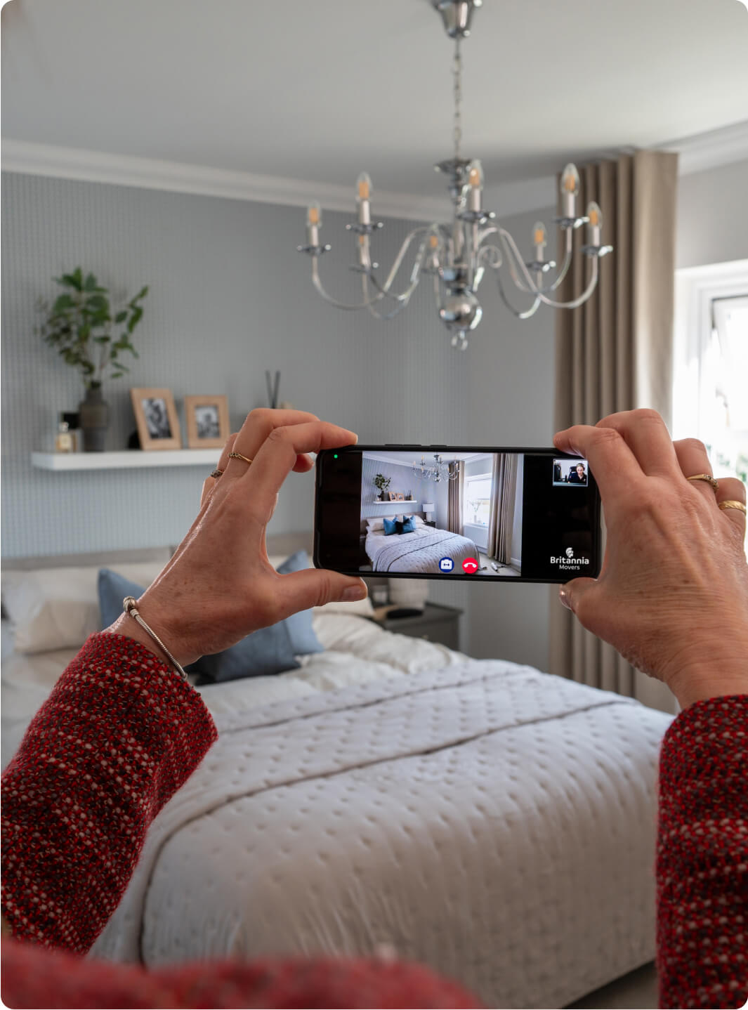 A person is using a smartphone to take a photo of a neatly made bed in a stylishly decorated bedroom. The room has a decorative chandelier, framed pictures on a shelf, and a potted plant beside the bed. The smartphone screen shows the bedroom image being captured.