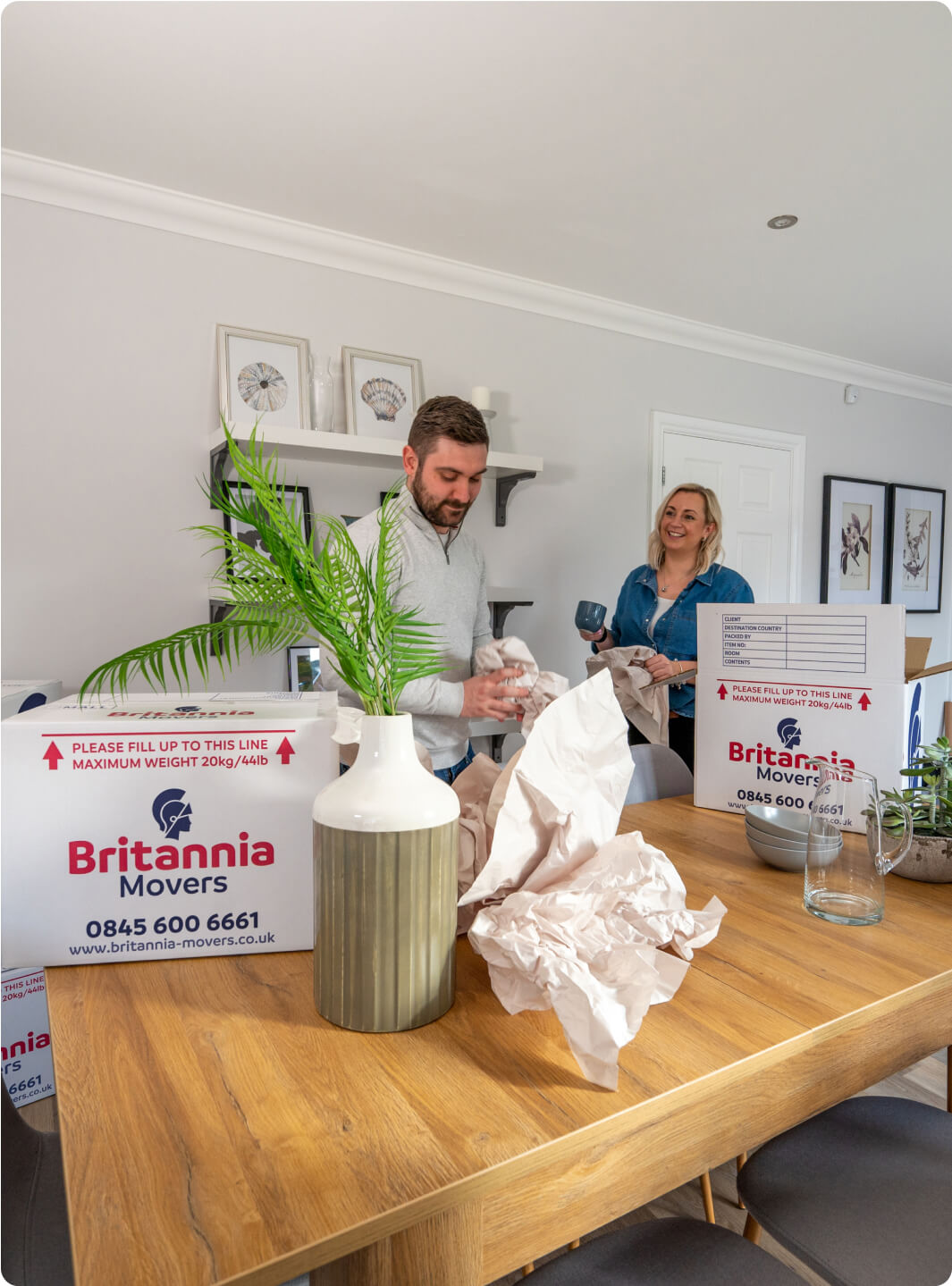 A man and woman are unpacking items from moving boxes labeled "Britannia Movers" in a bright, modern kitchen or dining area. They are surrounded by packing paper and household items, including a large vase with green foliage on the wooden table.