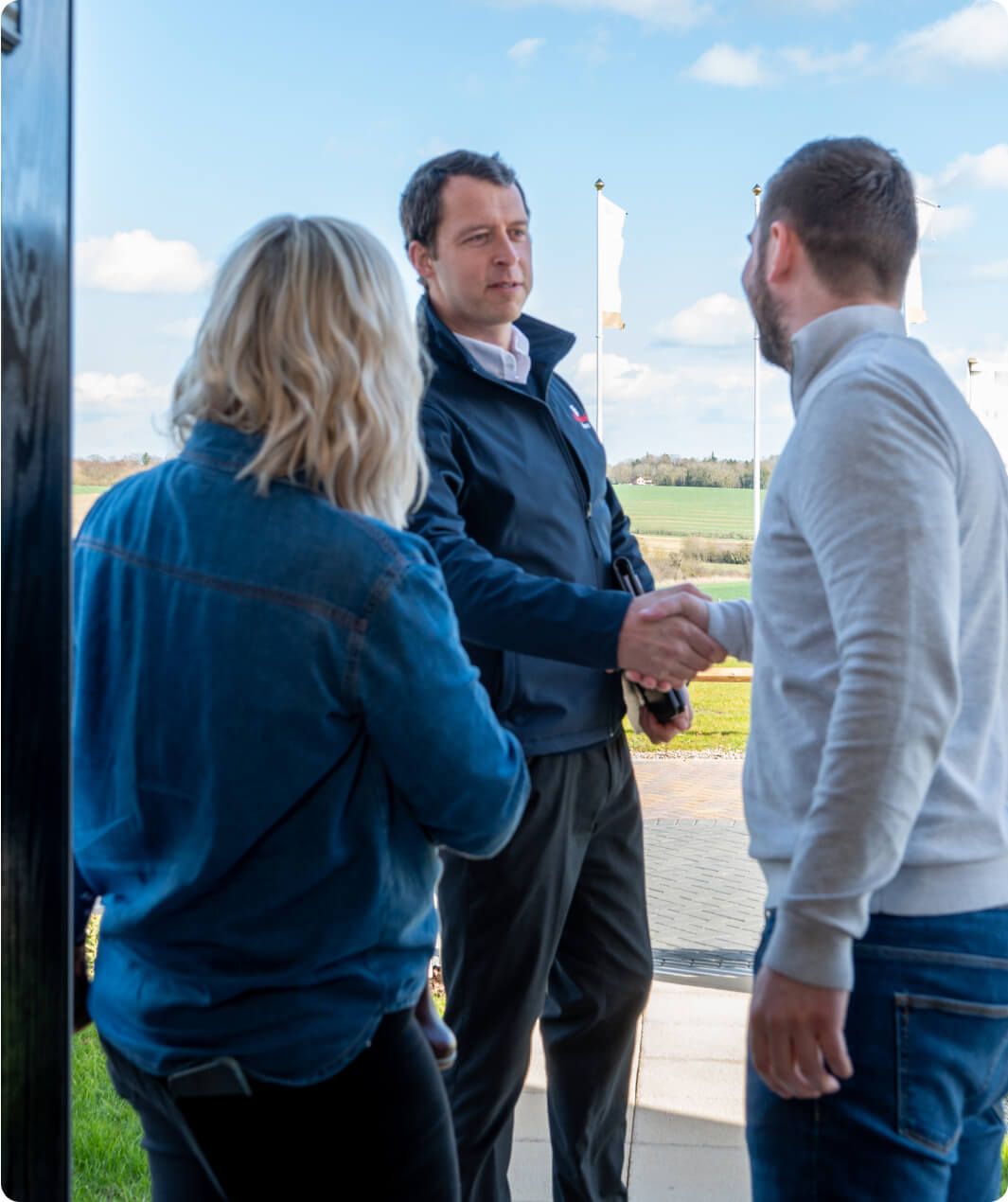 Three people standing and conversing outdoors. A man in a black jacket is shaking hands with a man in a light sweater, while a woman with blonde hair, wearing a denim jacket, stands nearby. The background features a clear sky and some flags.