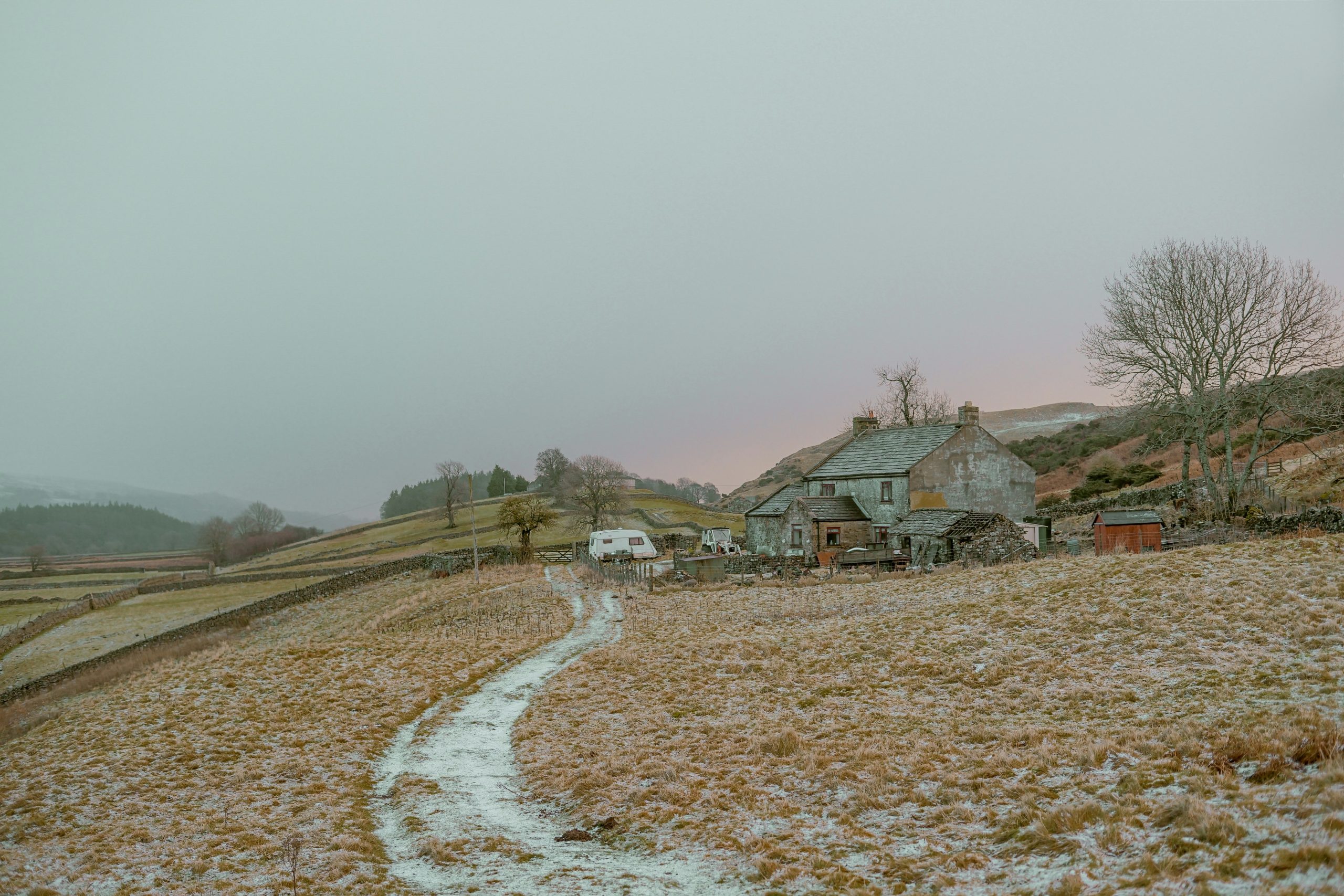 A serene winter landscape featuring a rustic stone house with a snow-dusted path leading up to it. The surrounding fields are lightly covered with snow, and bare trees stand amid the rolling hills. A caravan is parked nearby, enhancing the peaceful, rural scene.