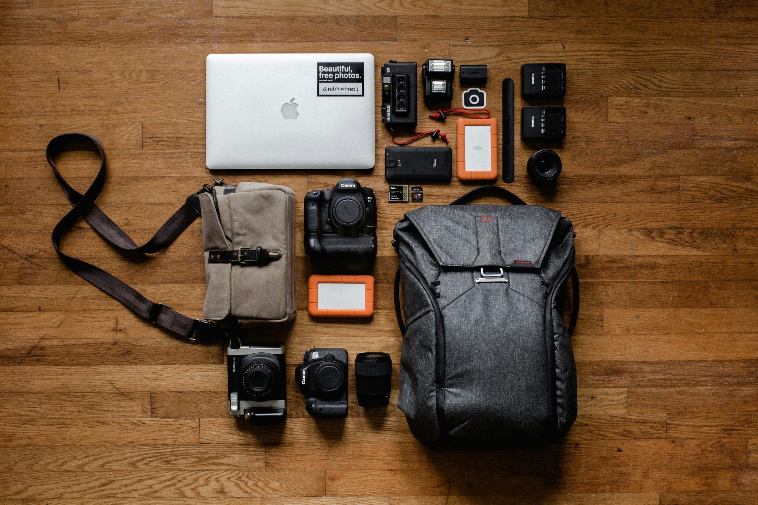 An array of photography equipment laid out on a wooden floor. Items include a laptop with a "Beautiful things photos" sticker, camera bodies, lenses, battery packs, memory cards, a grey backpack, a small beige shoulder bag, and orange external hard drives.