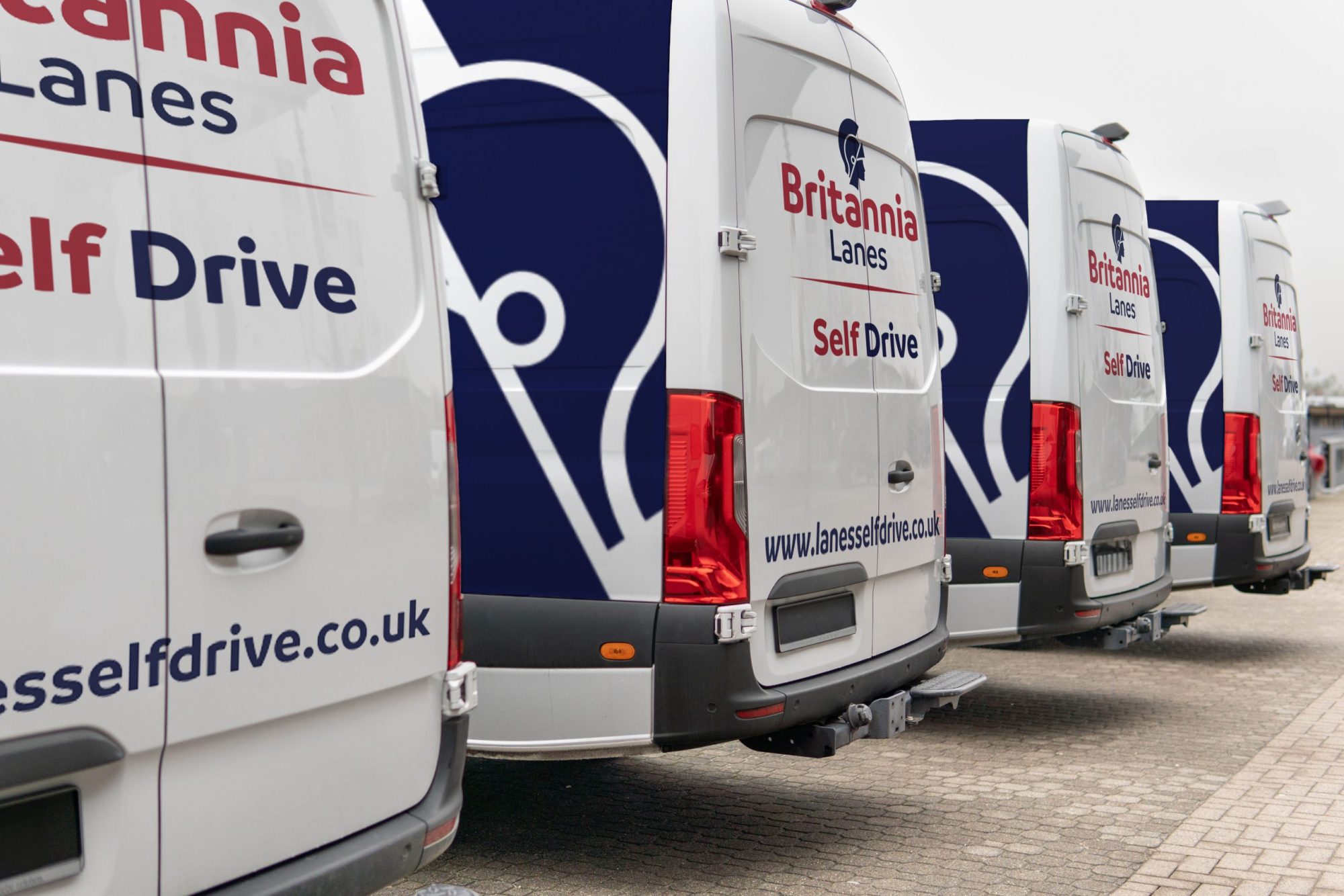 Four white vans are parked in a row, displaying the branding "Britannia Lanes Self Drive" along with a website address "lanesselfdrive.co.uk" in blue and red text on the rear and side. The vans are aligned on a paved surface.