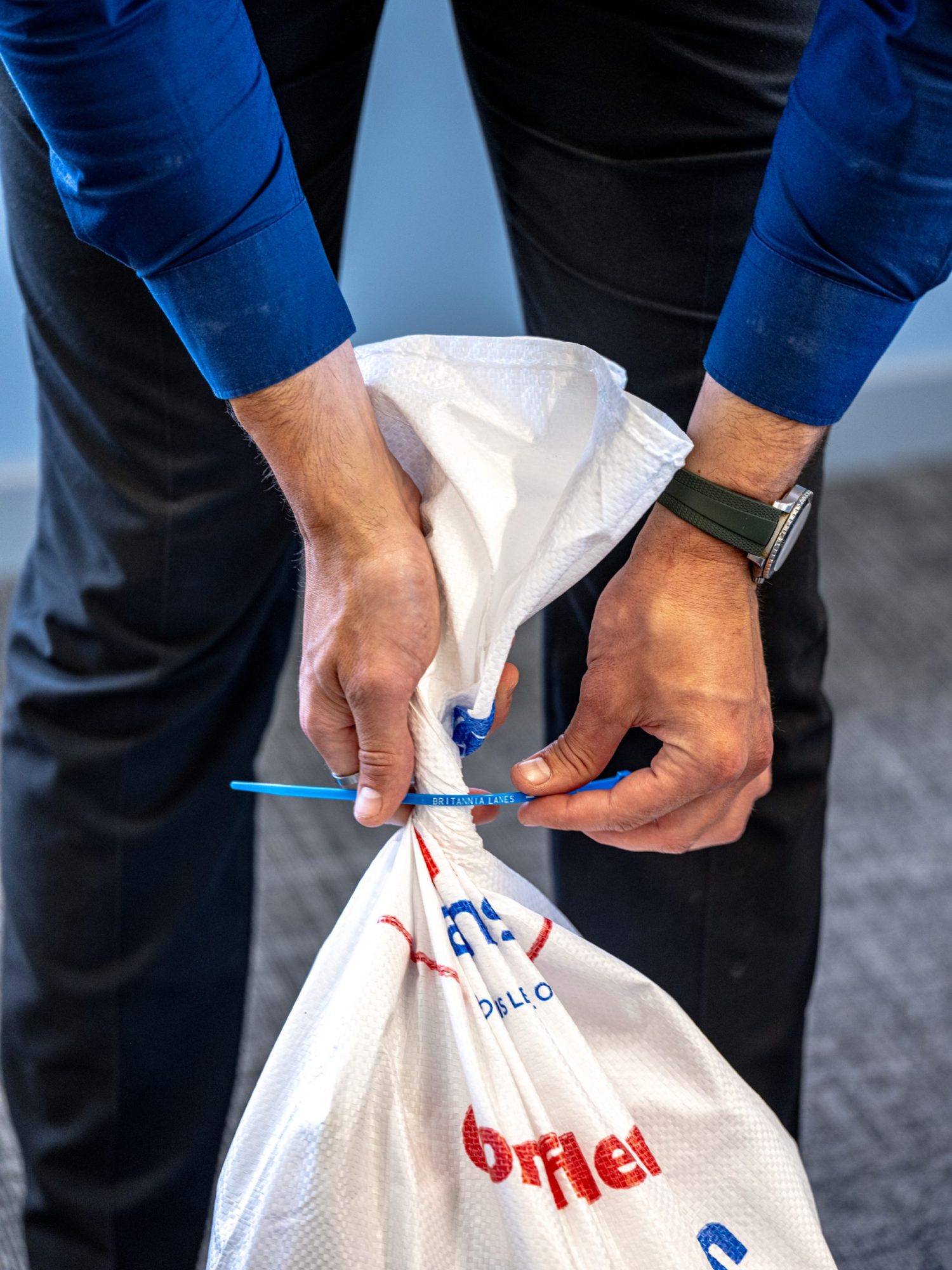 A person wearing a blue shirt and black pants tightens a white plastic bag with a blue zip tie. The bag has red and blue text printed on it. The individual is standing on a grey carpeted floor.