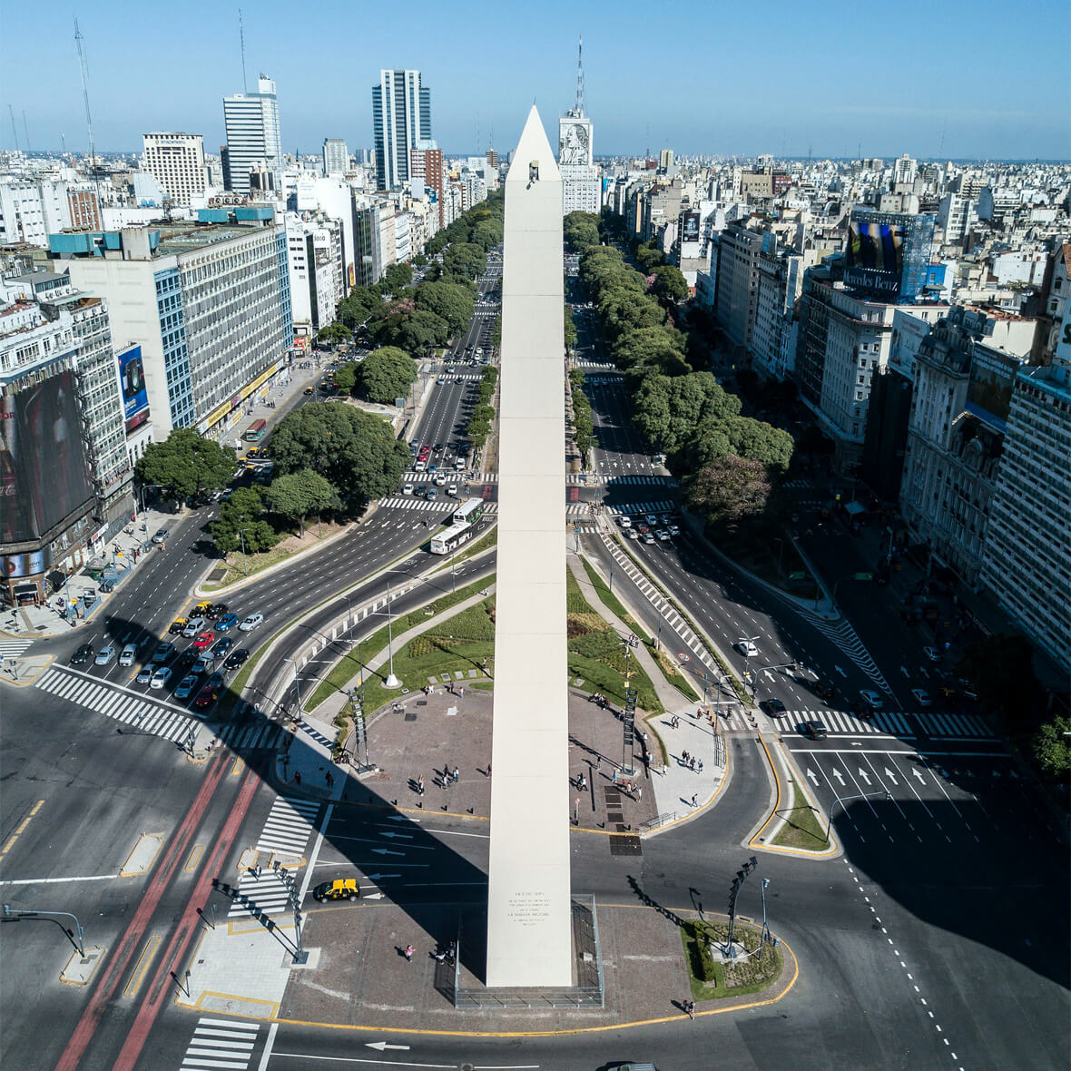 Aerial view of the Obelisk of Buenos Aires situated at the intersection of two major avenues, surrounded by traffic lanes and greenery. Tall buildings line both sides of the avenues, extending into the distance under a clear blue sky.