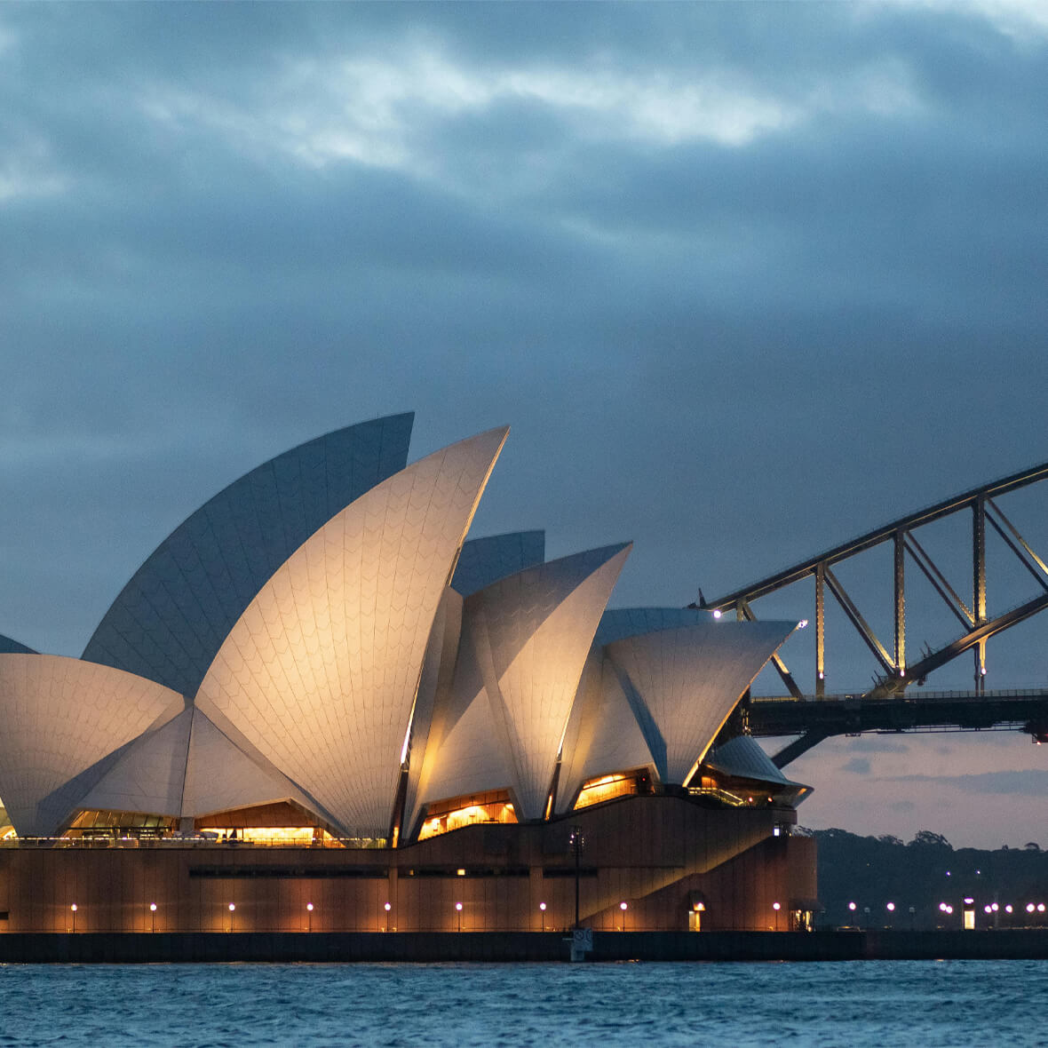 The image shows the Sydney Opera House illuminated at dusk, with its iconic sail-like structures. In the background, part of the Sydney Harbour Bridge is visible, extending toward the right side of the image. The sky is cloudy, and the water in the foreground is calm.