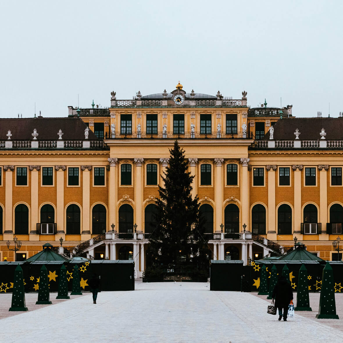 A grand yellow palace with rows of windows and an ornate facade, centered by an imposing Christmas tree. Flanked by green booths decorated with yellow stars, people are walking on the snowy ground in front of the palace. The sky is overcast.