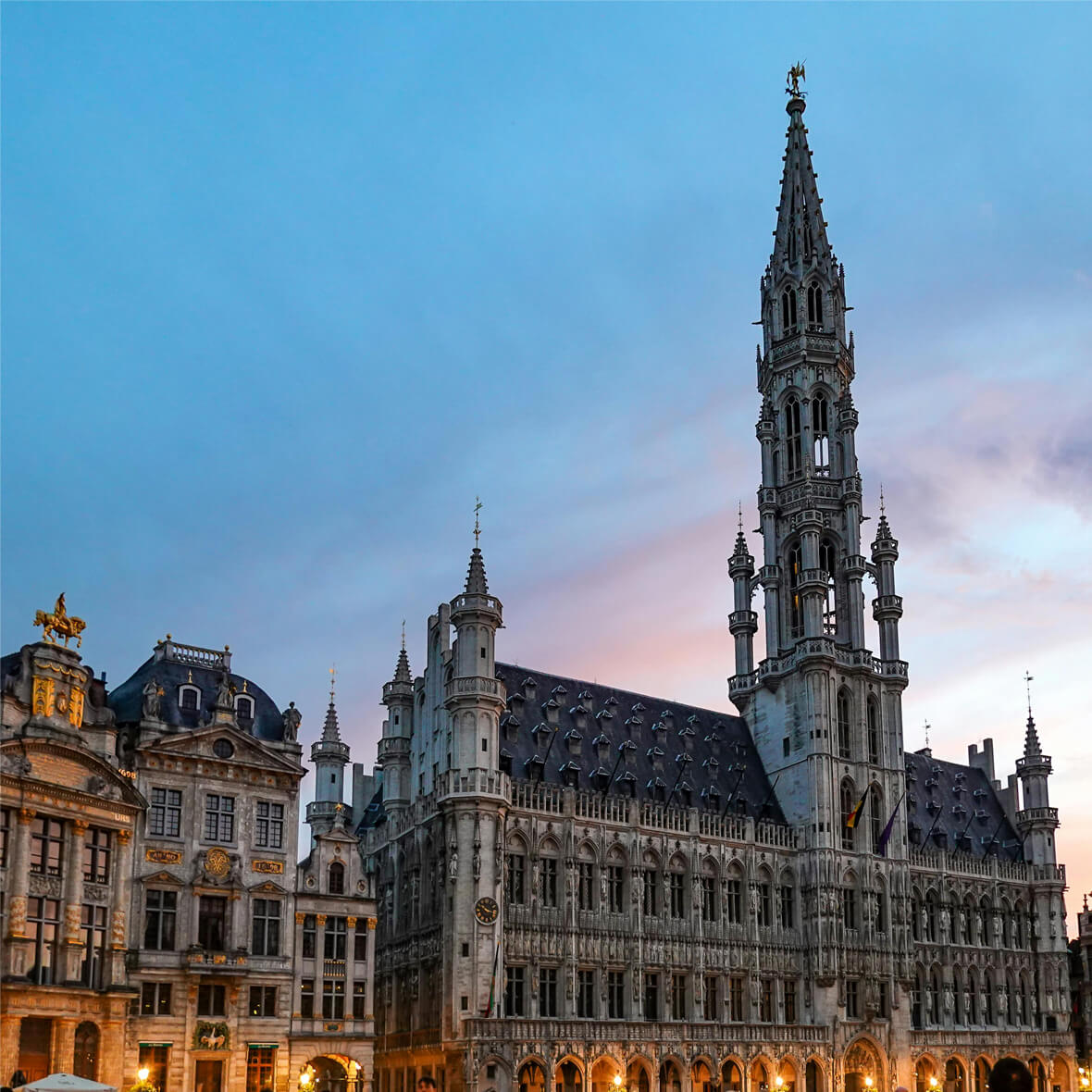 A majestic Gothic-style building, the Town Hall of Brussels, stands tall at dusk in the Grand Place. The sky is a gradient of blue to pink, and the intricate architectural details are highlighted by soft lighting, creating a picturesque scene.