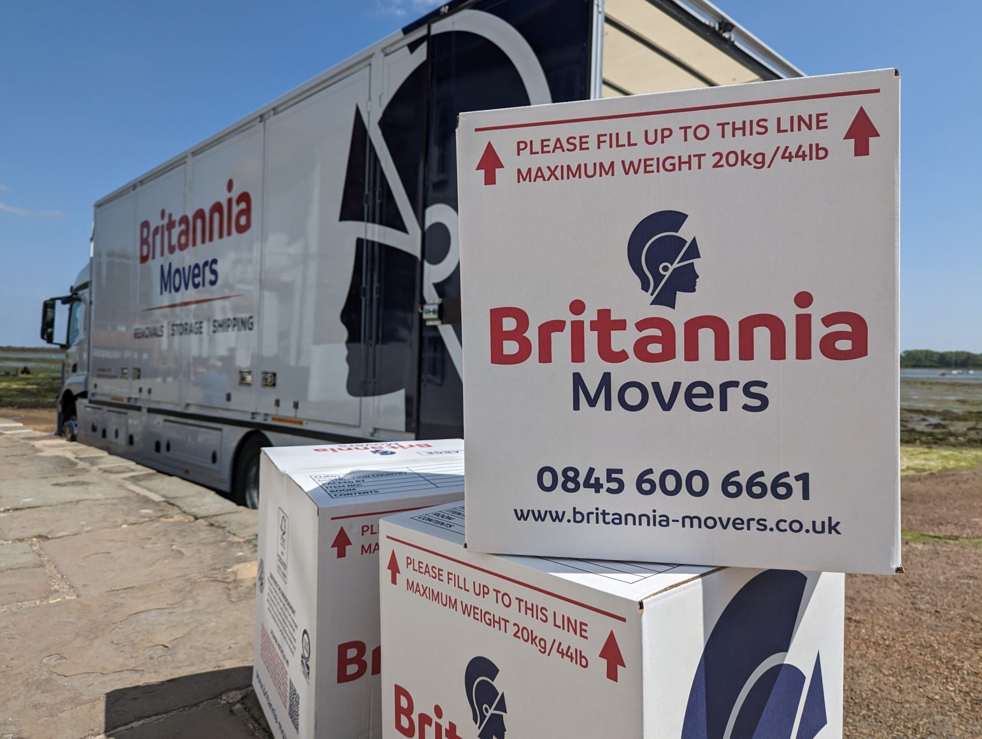 boxes-and-truck-on-the-beach A Britannia Movers truck is parked outdoors. In front of the truck, several boxes are stacked, displaying the Britannia Movers logo, contact number (0845 600 6661), and website (www.britannia-movers.co.uk). The boxes indicate a maximum weight limit of 20kg/44lb.