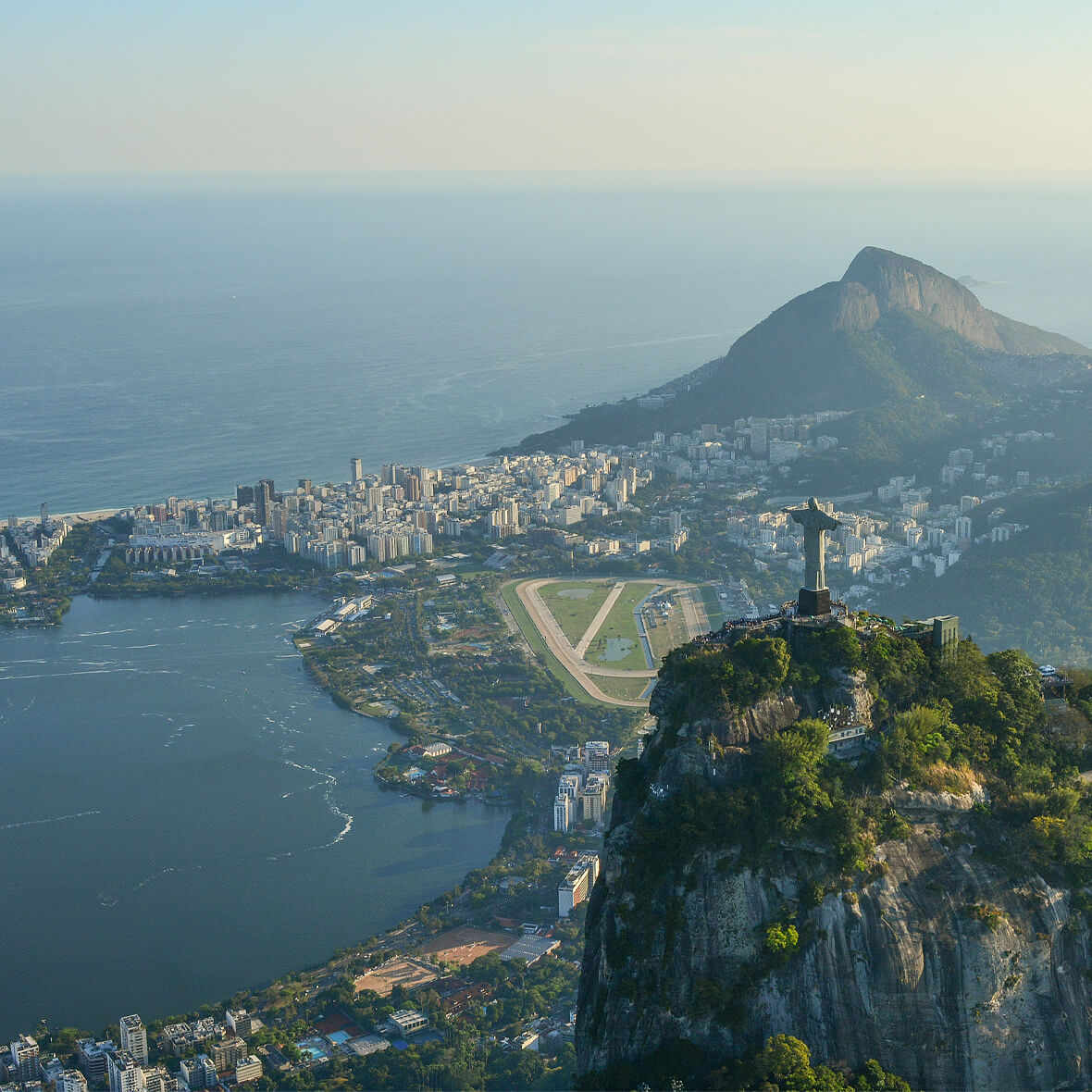 Aerial view of Rio de Janeiro, Brazil, featuring the iconic Christ the Redeemer statue atop Corcovado Mountain. The cityscape includes Guanabara Bay, an airstrip, and the surrounding buildings, with the Sugarloaf Mountain prominent in the background.