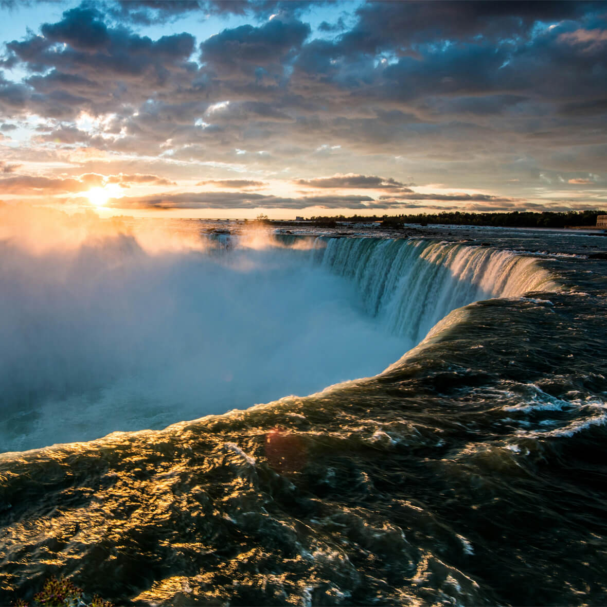 A stunning view of a large waterfall with water cascading over the edge, creating mist. The sun sets in the background, casting a warm golden glow over the scene. The sky is partly cloudy, and the horizon is lit with soft sunlight.