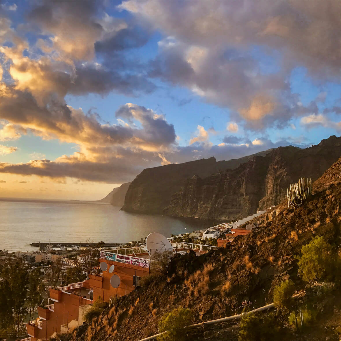 A coastal town nestled against steep cliffs at sunset. The sky is dotted with clouds, illuminated in hues of gold and orange. The sea is calm, with the coastline extending into the distance. Buildings are scattered along the hillside, with cacti and vegetation in the foreground.