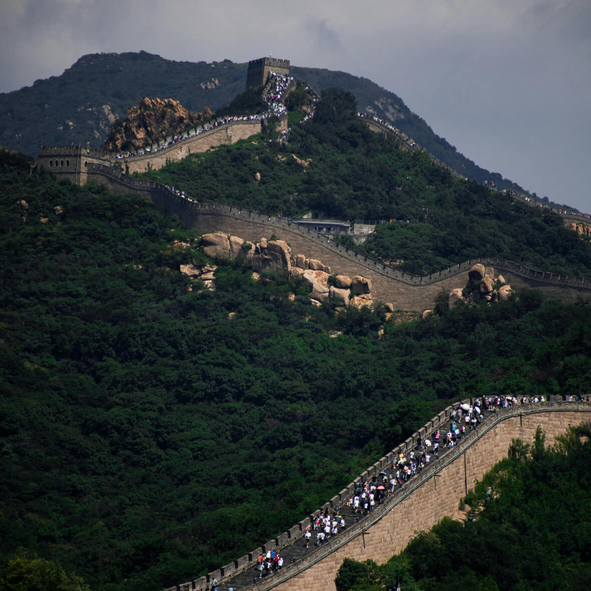 A section of the Great Wall of China stretches across green, forested mountains. Tourists are walking along the wall, some reaching the watchtowers. The sky is overcast, adding a misty atmosphere to the scenic landscape.