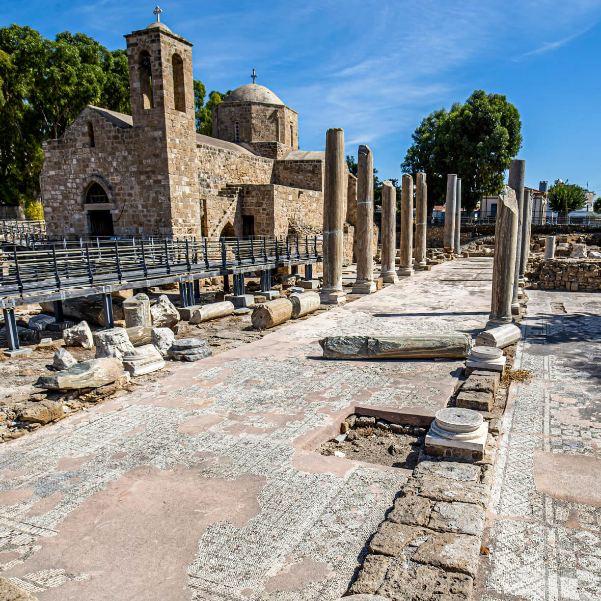 Ancient ruins with stone columns and detailed mosaics in the foreground. A historic stone church with a cross on the roof stands nearby. Bright blue sky and greenery in the background. Stone pathways and platforms surround the structures.