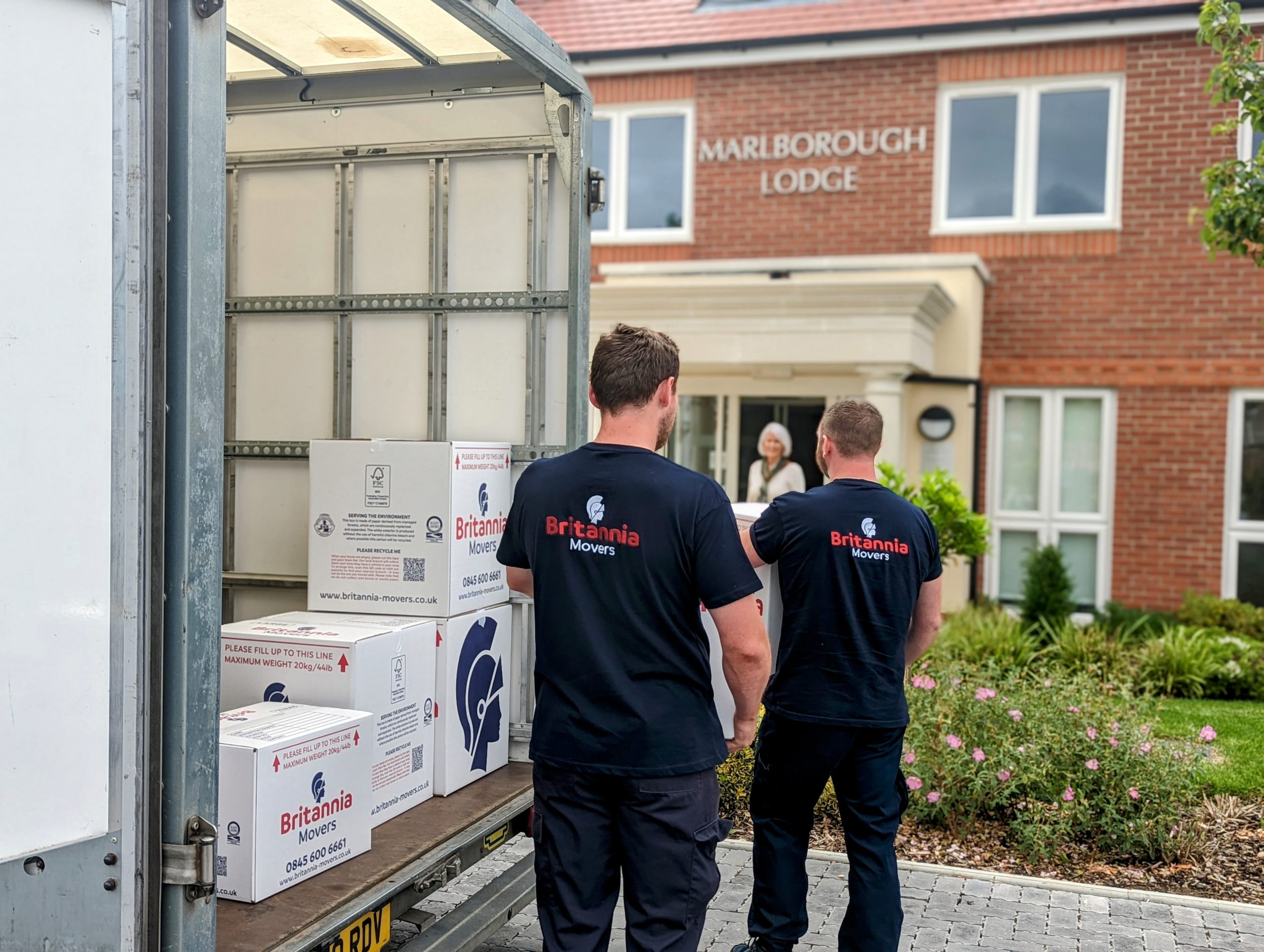 Two movers, wearing "Britannia Moves" shirts, carry boxes from a moving truck parked outside a building labeled "Marlborough Lodge." The truck's open back reveals packing boxes inside, while a flower bed is visible in the foreground.