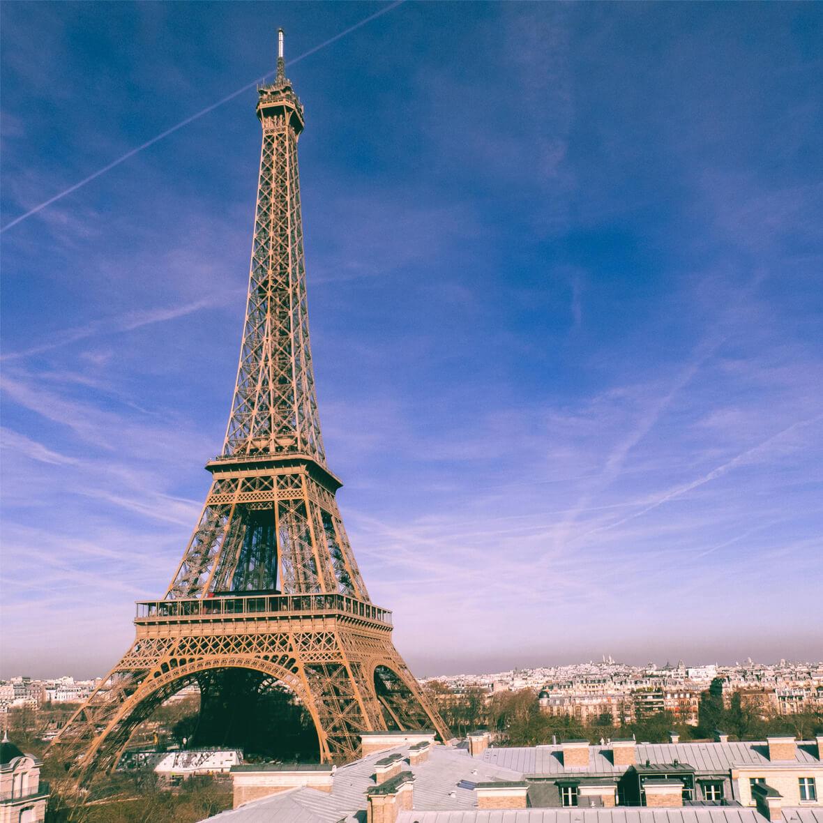 The Eiffel Tower under a clear blue sky with wispy clouds and airplane contrails. The iconic structure stands tall in the center of the image, with the city of Paris visible in the background. Rooftops of nearby buildings can be seen in the foreground.