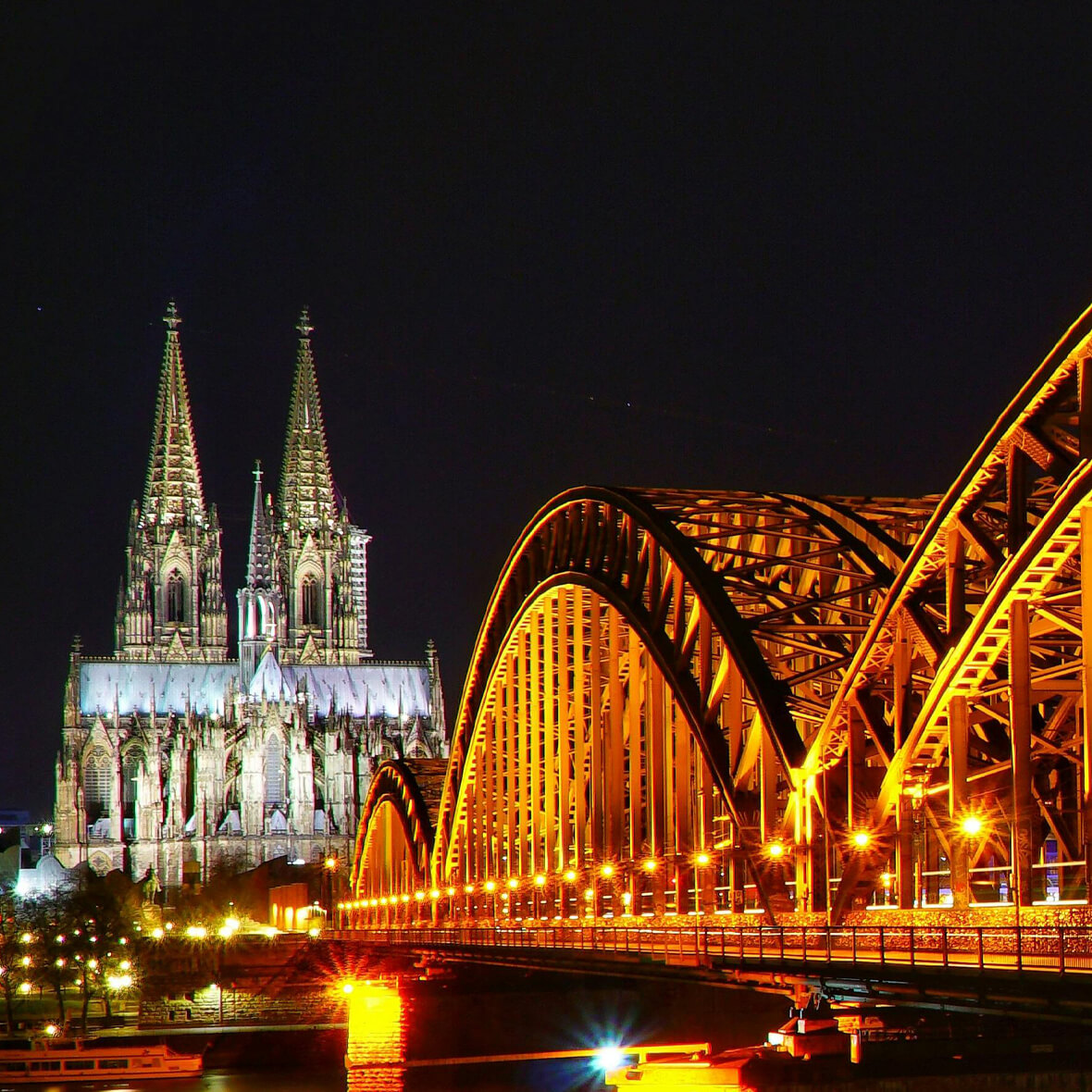 A nighttime view of a brightly-lit Gothic cathedral with twin spires, adjacent to an illuminated arched bridge spanning a river. The scene is set against a dark sky, with various boats visible on the water below.