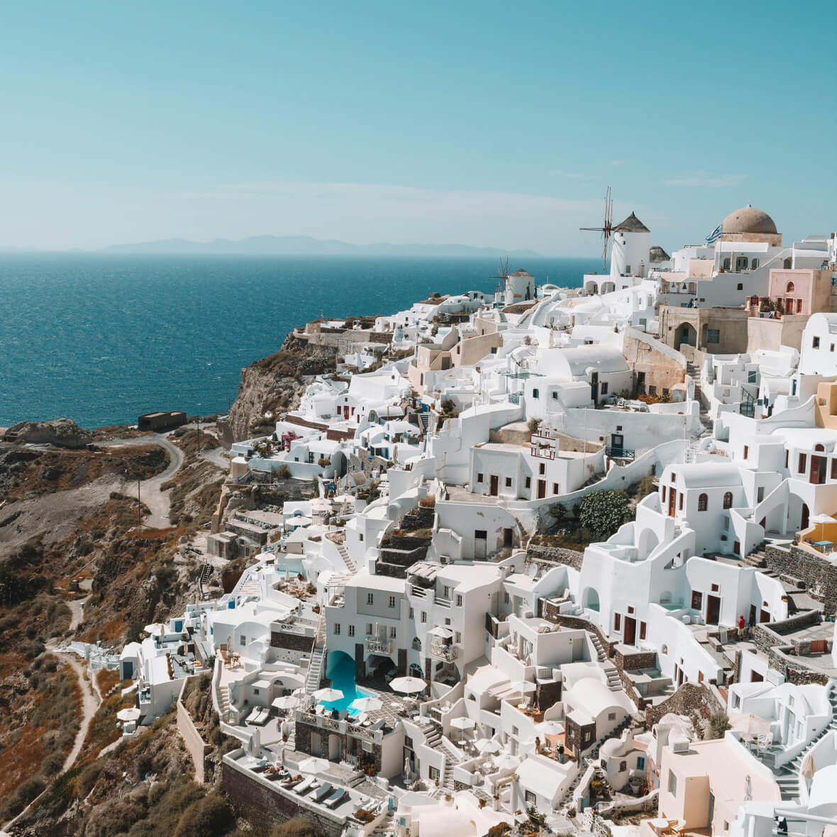 A scenic view of a coastal town in Santorini, Greece. The picture features white-washed buildings with blue roofs cascading down a hillside, overlooking the deep blue sea. A windmill and a domed structure are visible in the background under a clear sky.