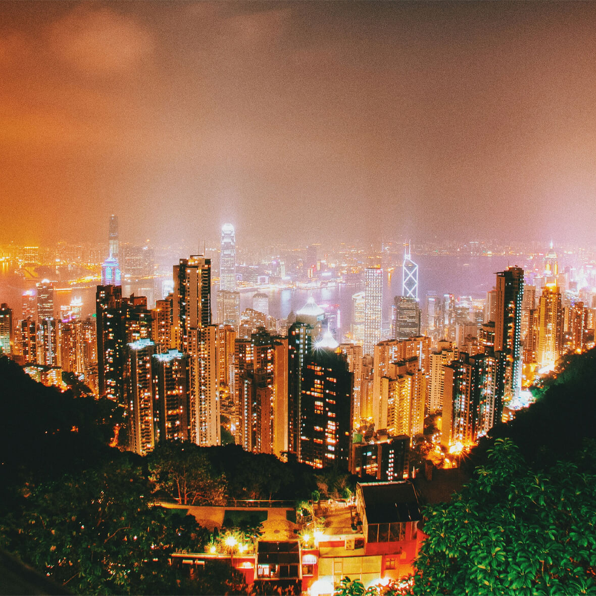 A vibrant cityscape at night with tall skyscrapers illuminated with colorful lights. The skyline is dotted with numerous buildings, with some of the tallest structures distinctly bright. A hazy glow surrounds the city against a backdrop of a dark sky.