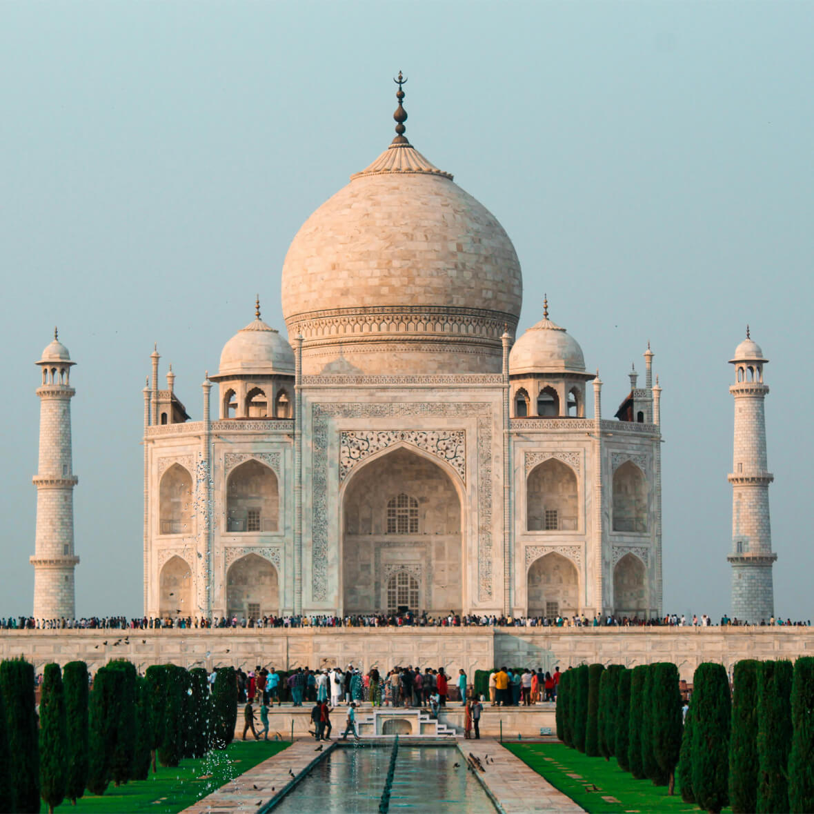 A large white marble mausoleum with a massive dome and minarets at each corner stands majestically against a clear sky. It is surrounded by manicured lawns and a reflecting pool, with crowds of people gathered near its base.