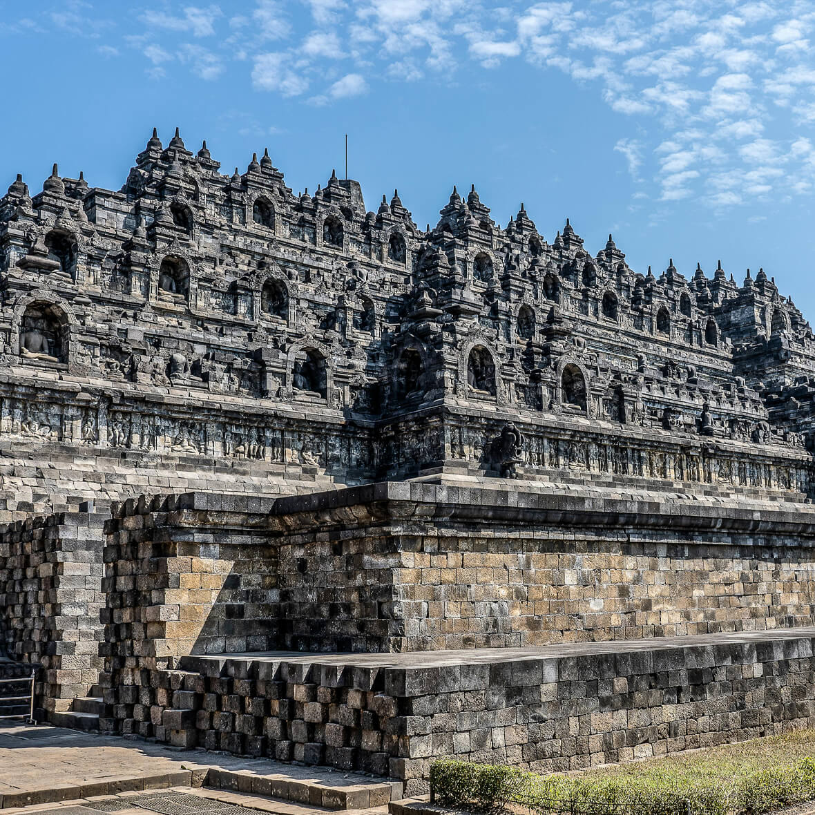 A large, ancient stone temple with intricate carvings and multiple spires stands under a clear blue sky. The structure exhibits significant gray detailing and is built on a multi-tiered platform, with a foreground of trimmed grass and a stone pathway.