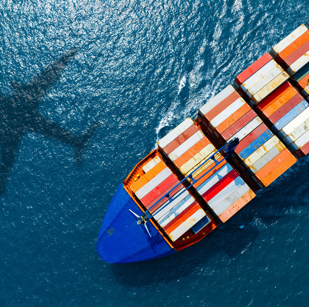 Aerial view of a cargo ship with colorful containers sailing on the ocean. The shadow of an airplane can be seen on the water's surface, suggesting the plane is flying overhead.