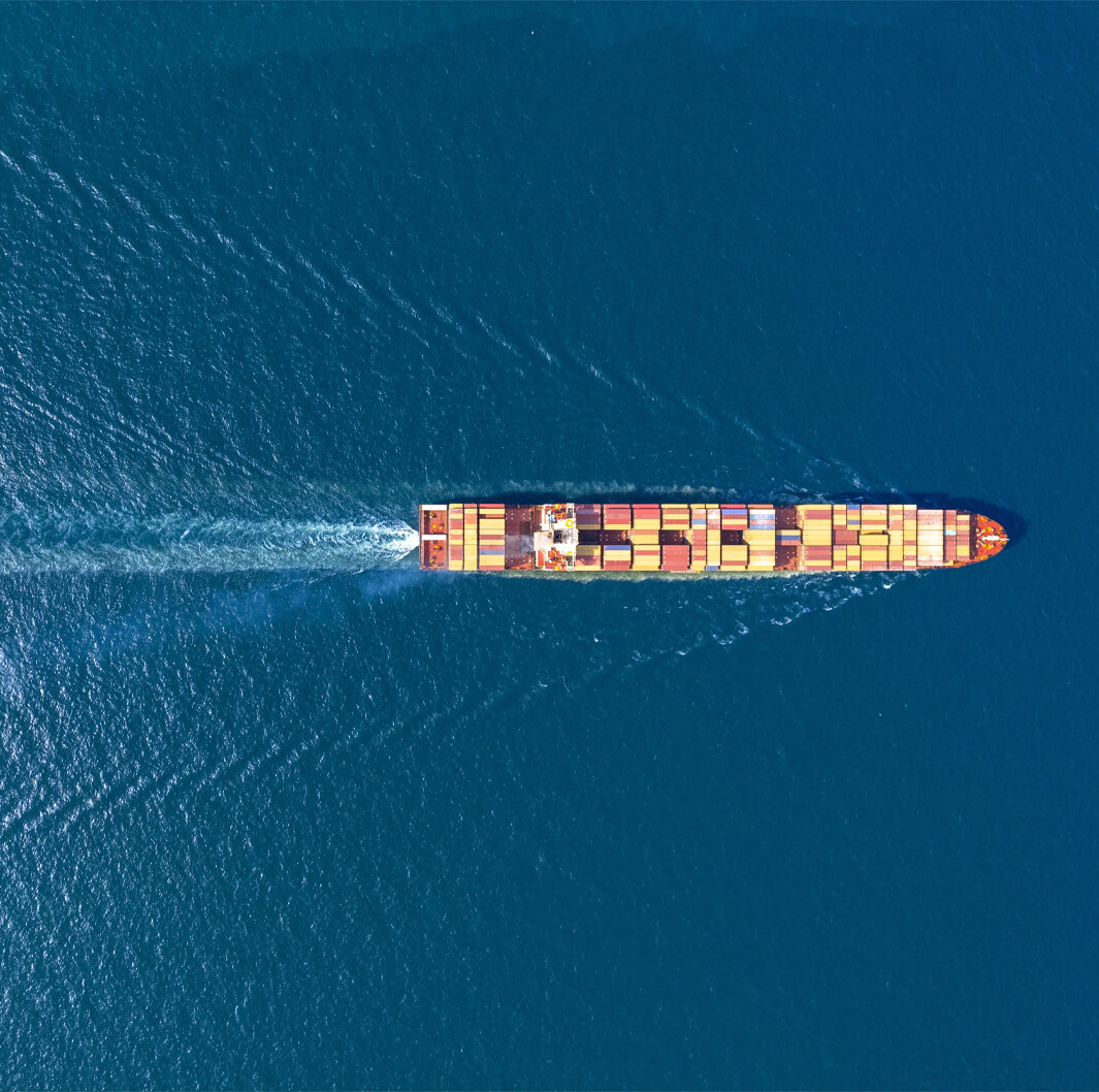 An overhead view of a large cargo ship sailing through deep blue ocean waters. The vessel is carrying numerous colorful containers arranged neatly along its deck. The ship creates a white wake as it moves forward, cutting through the serene sea.