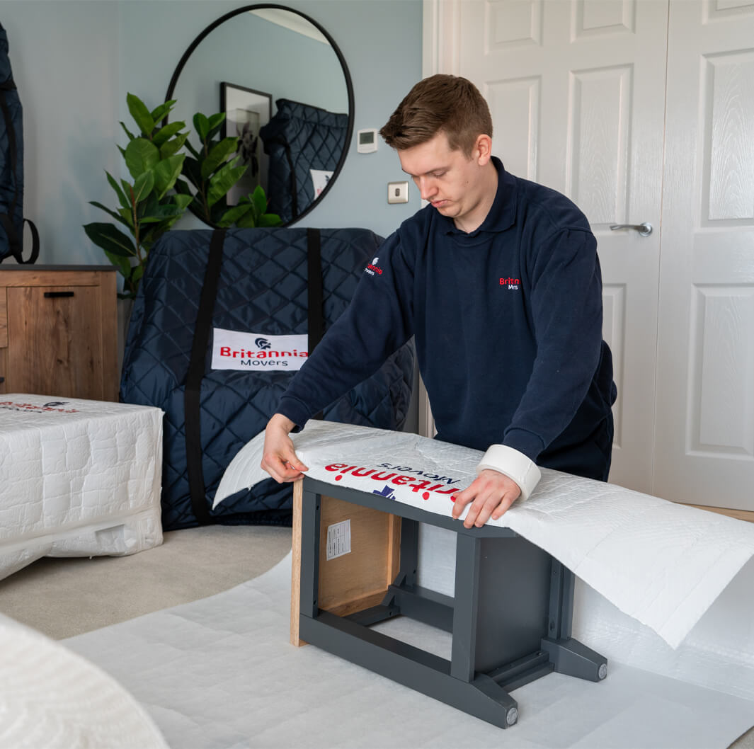 A person in a dark uniform wraps a piece of furniture with a white protective cover inside a room. Behind them, there are other wrapped items and green plants near a wooden dresser. A logo on the cover reads "Britannia Movers.