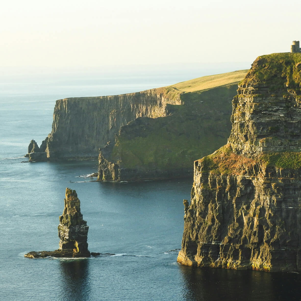 The image shows the Cliffs of Moher along the coastline in Ireland. The sheer rock faces rise dramatically from the Atlantic Ocean. A solitary rock formation stands prominently in the water just off the coast, and the landscape is bathed in soft golden light.