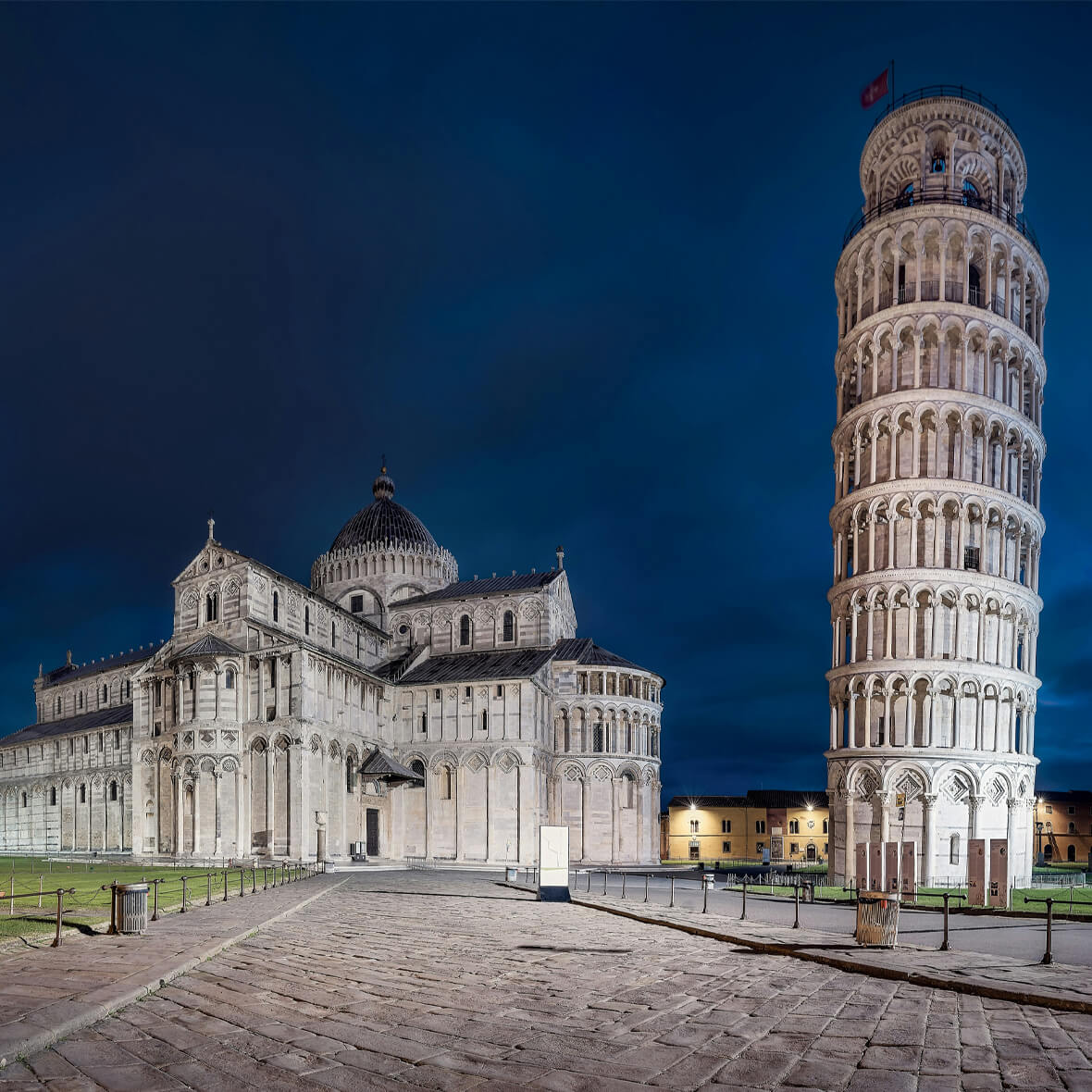 Night view of the Leaning Tower of Pisa and the adjacent cathedral. The tower, lit softly, tilts to the right, while the cathedral showcases Romanesque architecture. Cobblestone paths and a small fence are also visible in the foreground.