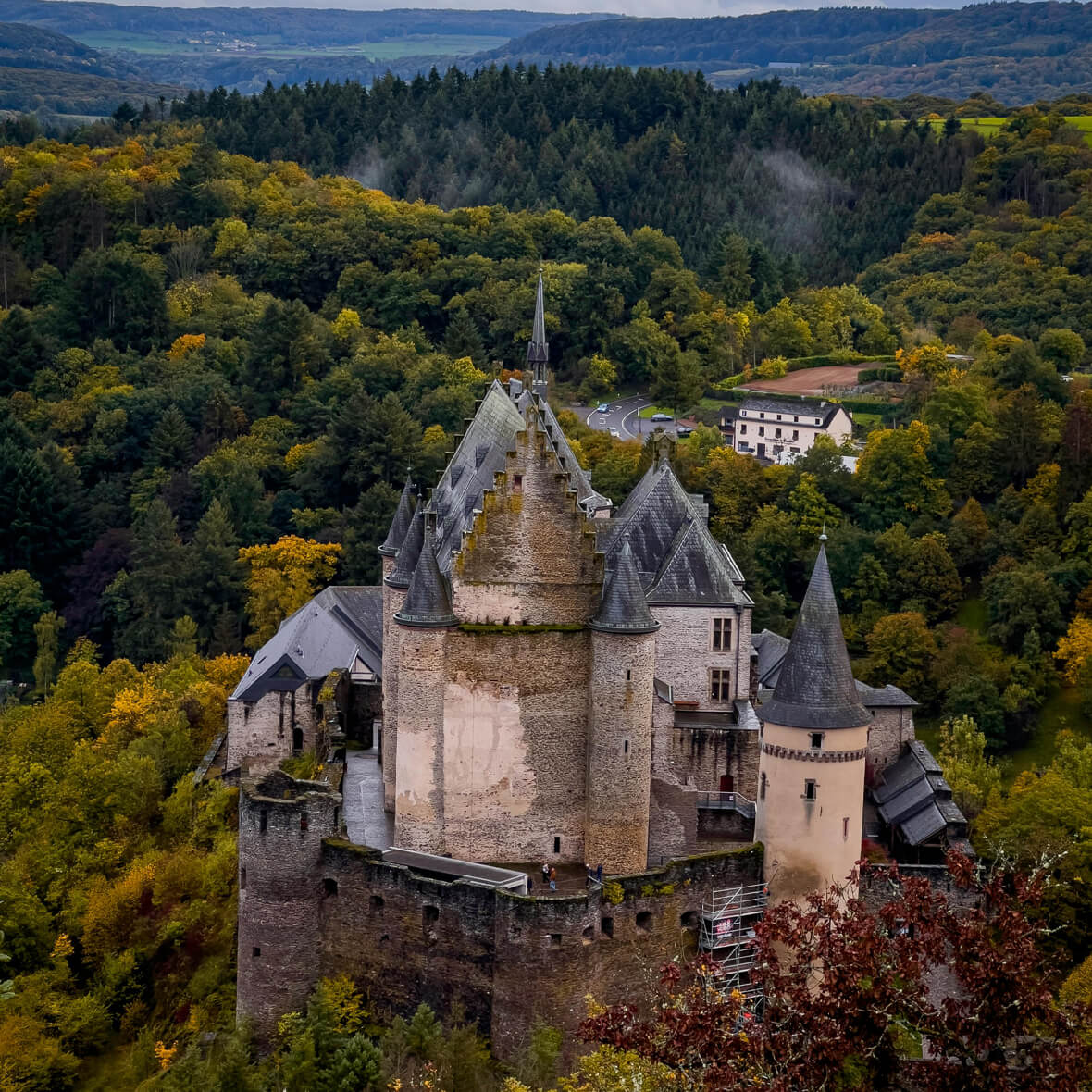 A medieval stone castle with multiple towers and a high central spire is nestled in a dense forest with autumn foliage. Rolling hills and a distant valley are visible in the background. A nearby white building is partially hidden among the trees.