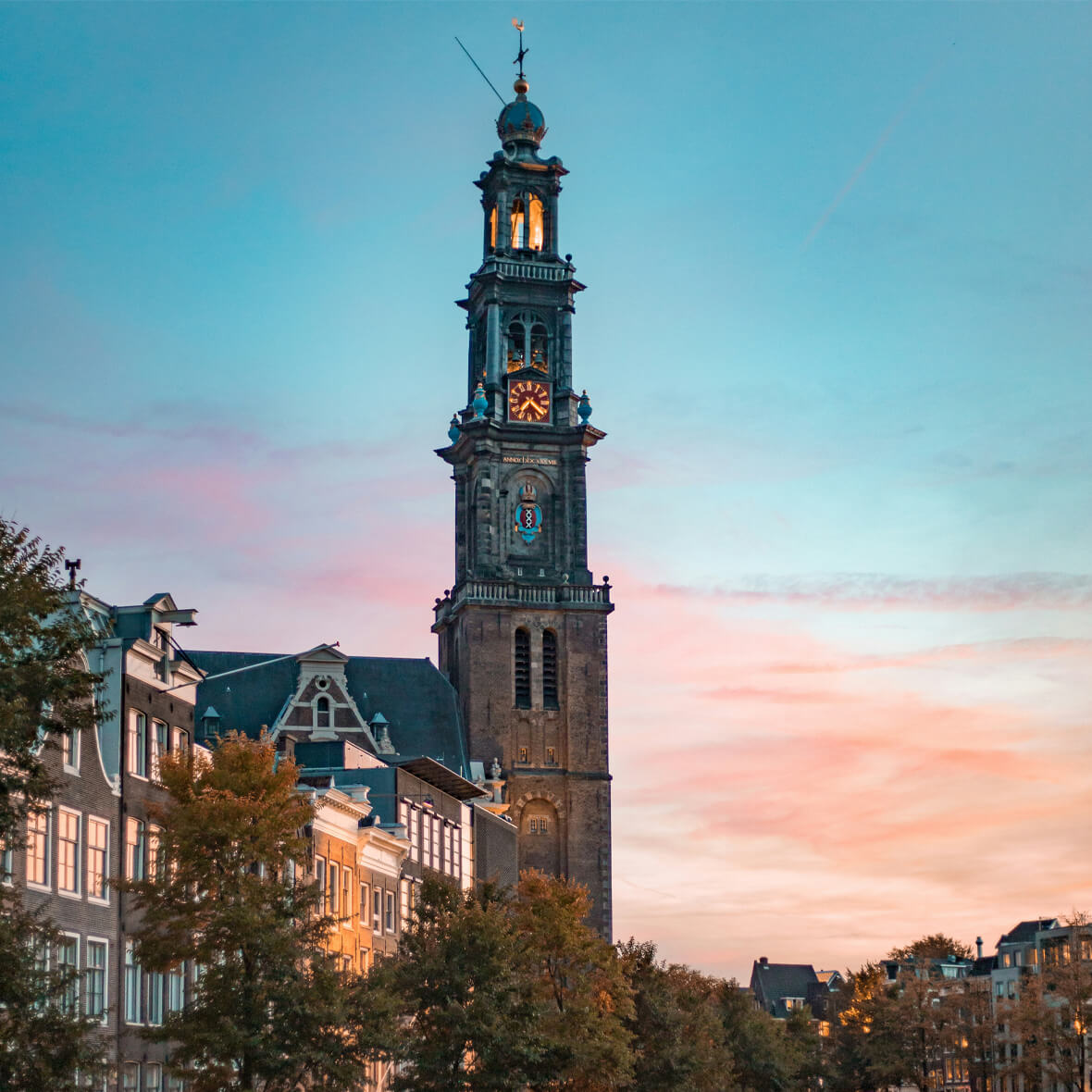 A tall, historic clock tower stands prominent against a backdrop of a pastel-colored sky during sunset. Surrounding the tower are traditional European-style buildings adorned with steep roofs and large windows, framed by lush green trees.